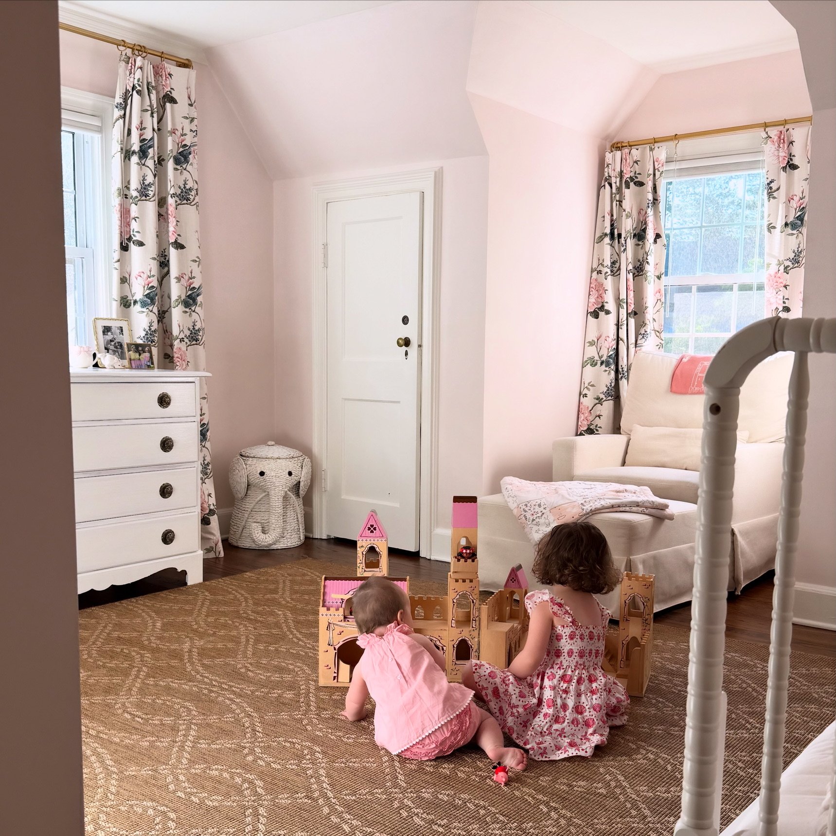 Two young children playing with a wooden castle toy on a brown patterned carpet in a bright room with pink walls, floral curtains, a white sofa with a pink pillow, and a white dresser with framed pictures and decorative items. The room has hardwood flooring and natural light coming through the windows.