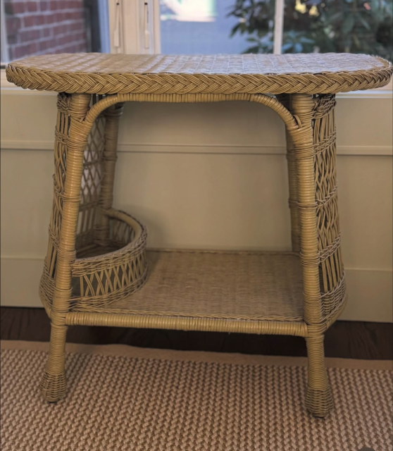 Wicker side table with a round top, two shelves, and curved supports, positioned on a beige rug in front of a window.