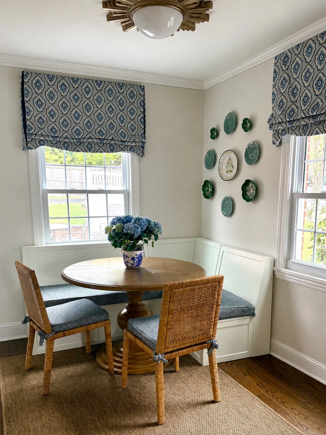 Dining nook with a round wooden table, two wicker chairs with cushions, built-in white corner bench, a vase with blue hydrangeas on the table, two windows with blue patterned valances, wall decoration with green plates and a central decorative plate, and hardwood flooring with a beige rug.