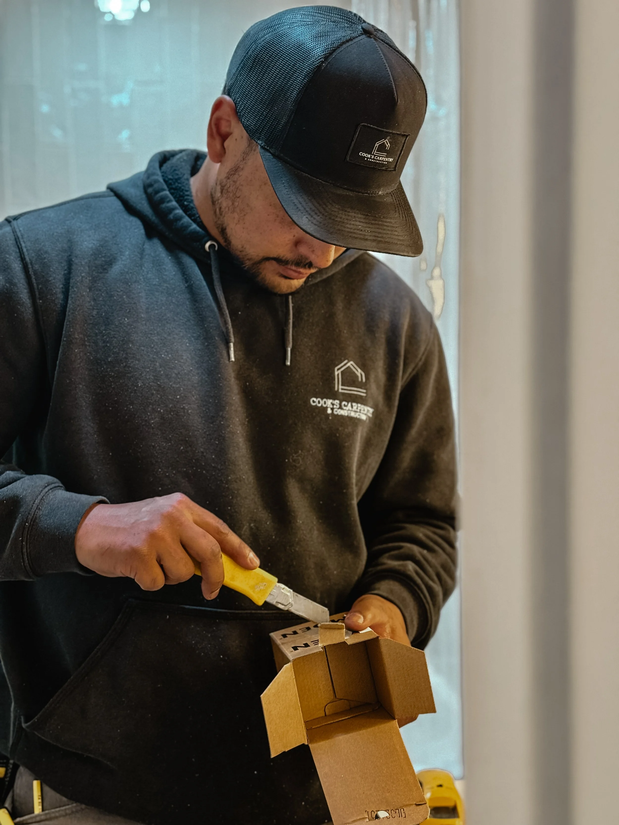 A man wearing a black cap and hoodie, with the hoodie displaying a logo that reads 'Cook's Carpentry & Construction', is working on a small wooden model or project. He is holding a yellow utility knife and appears to be carving or assembling the wooden piece.