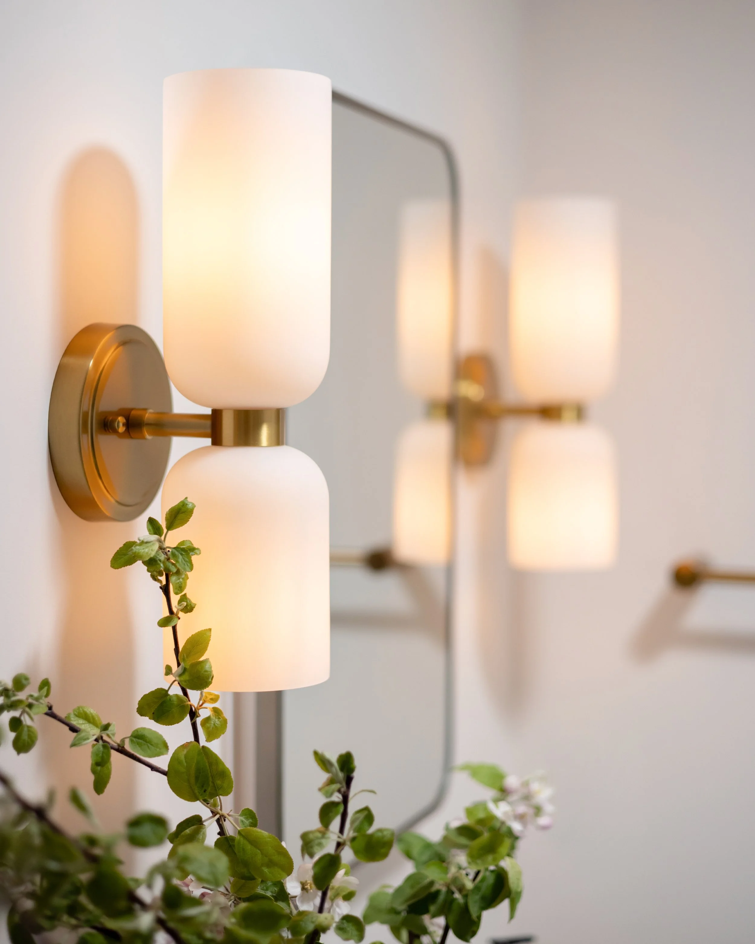 Close-up of a wall-mounted modern light fixture with a frosted glass shade and a brass arm, with green leafy plants in the foreground and other similar fixtures blurred in the background.