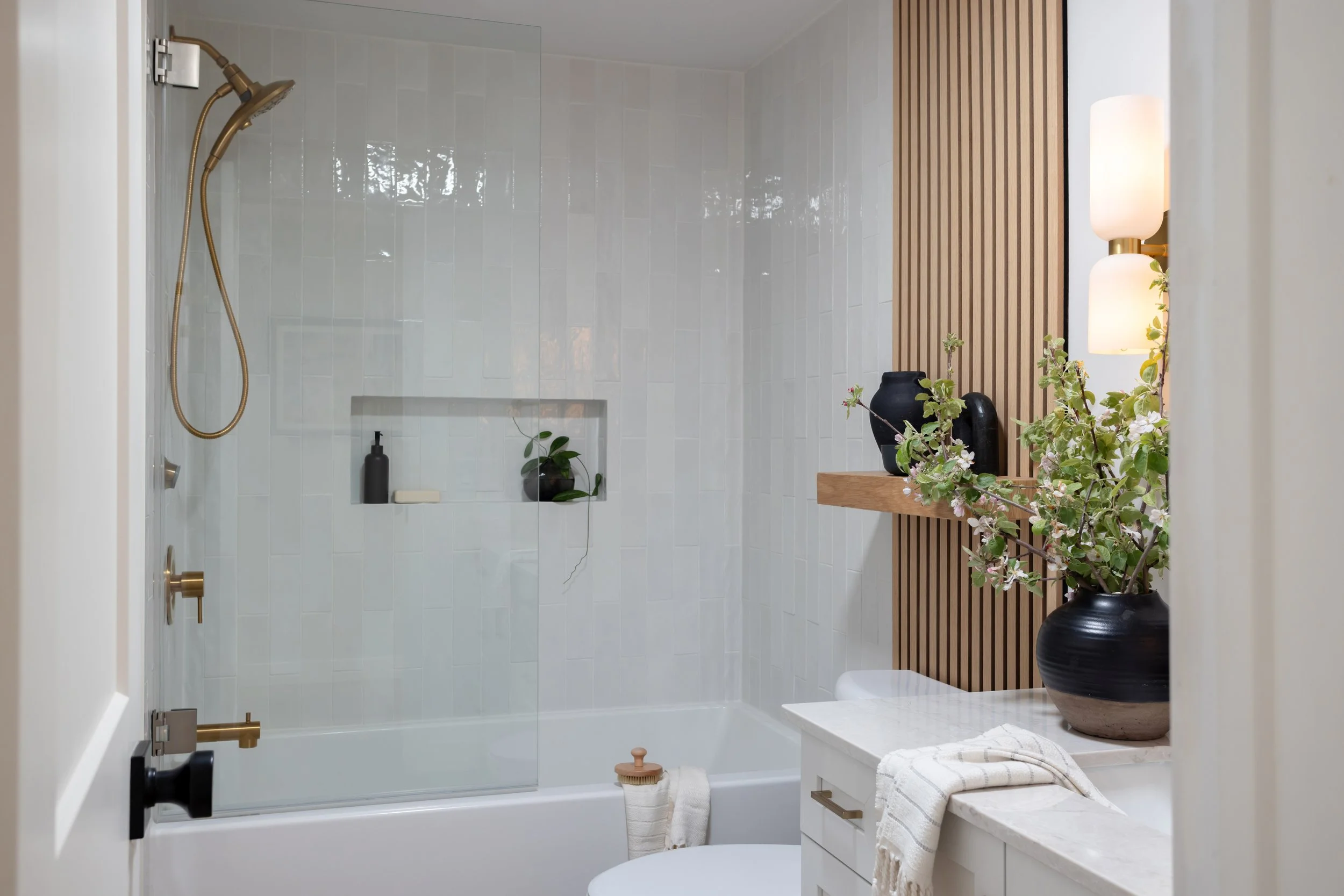Modern bathroom with a shower area featuring white tiles, a built-in niche with a plant and soap, a wooden slat accent wall, decorative vases, a lamp, and a marble countertop with a towel.