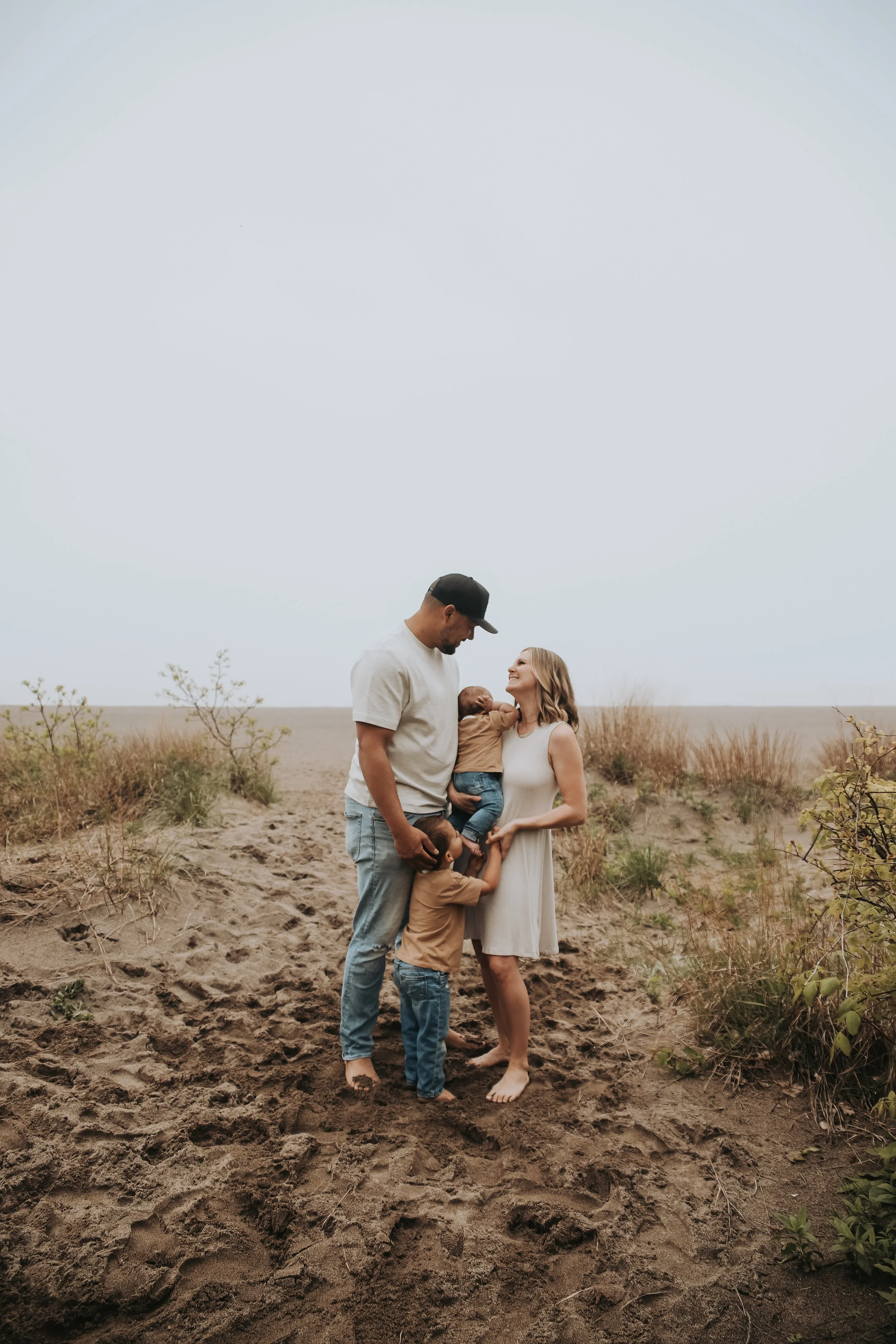 A family of four standing barefoot on sandy ground in an open field. The father, wearing a black cap and white T-shirt, looks down at his young son who is hugging his legs. The mother, wearing a white dress, is smiling and looking at the father. They are holding their newborn baby, who is wrapped in a blanket. The sky is overcast.