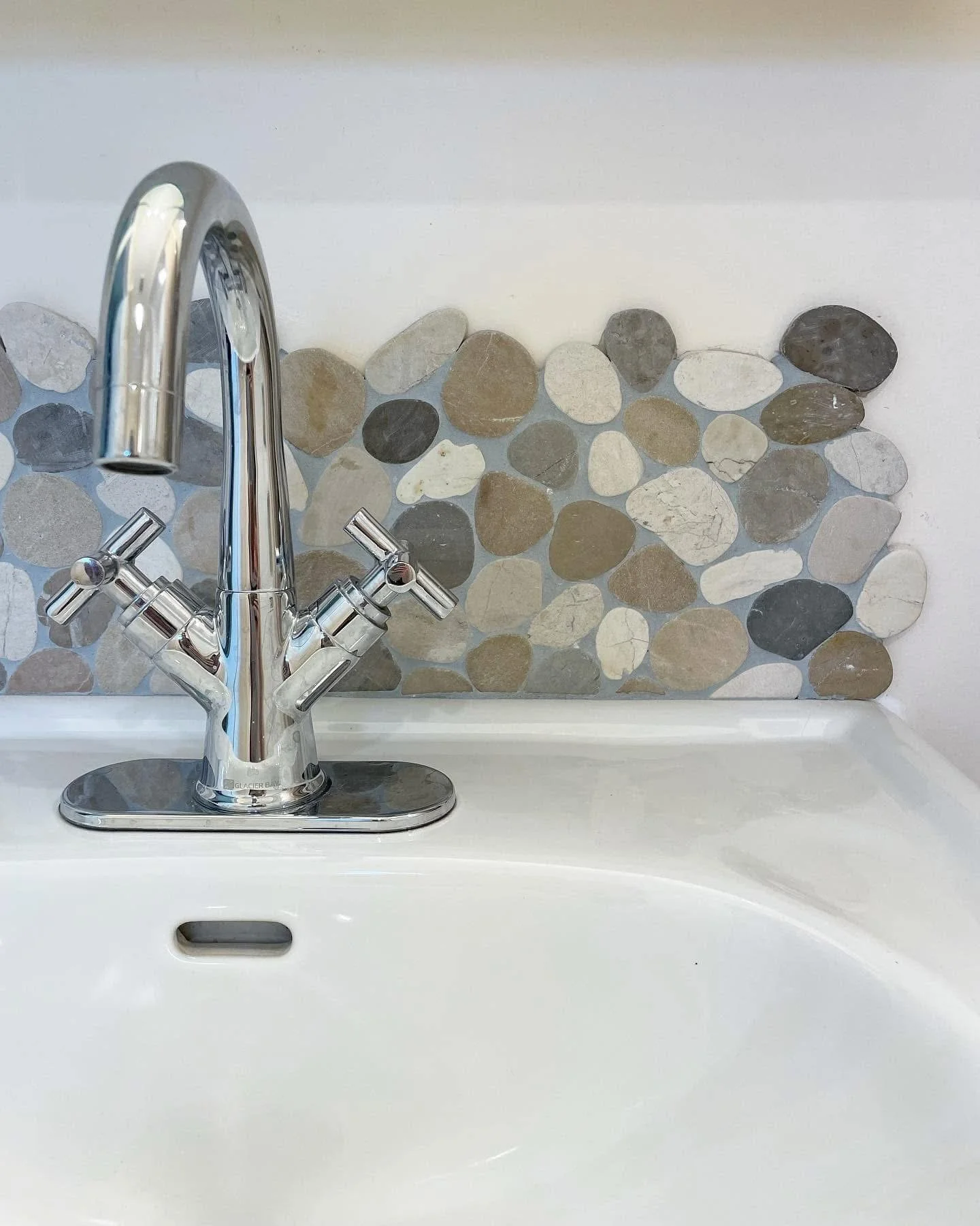 Close-up of a modern chrome kitchen faucet with dual handles, installed over a white sink with a pebble tile backsplash in shades of gray, beige, and white.