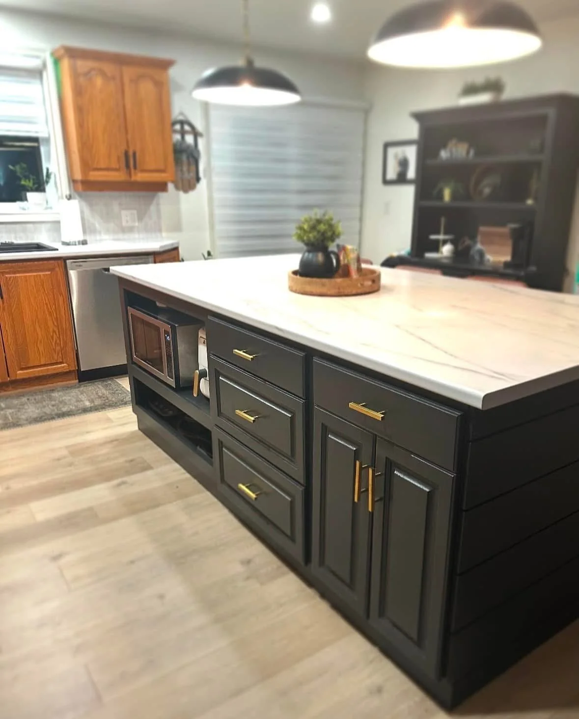 A modern kitchen with black and wooden cabinets, a white countertop island with gold handles, a microwave, pendant lighting, and a small decorative plant on a tray.