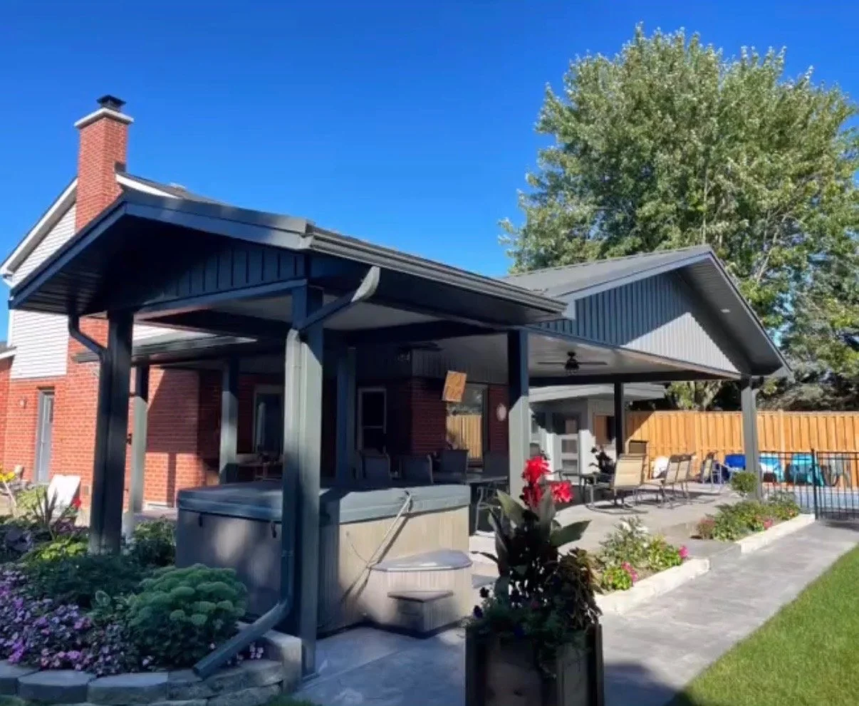Backyard patio with seating area, hot tub, shaded cover, potted plants, and a wooden fence, with a brick house and large tree in the background under a clear blue sky.