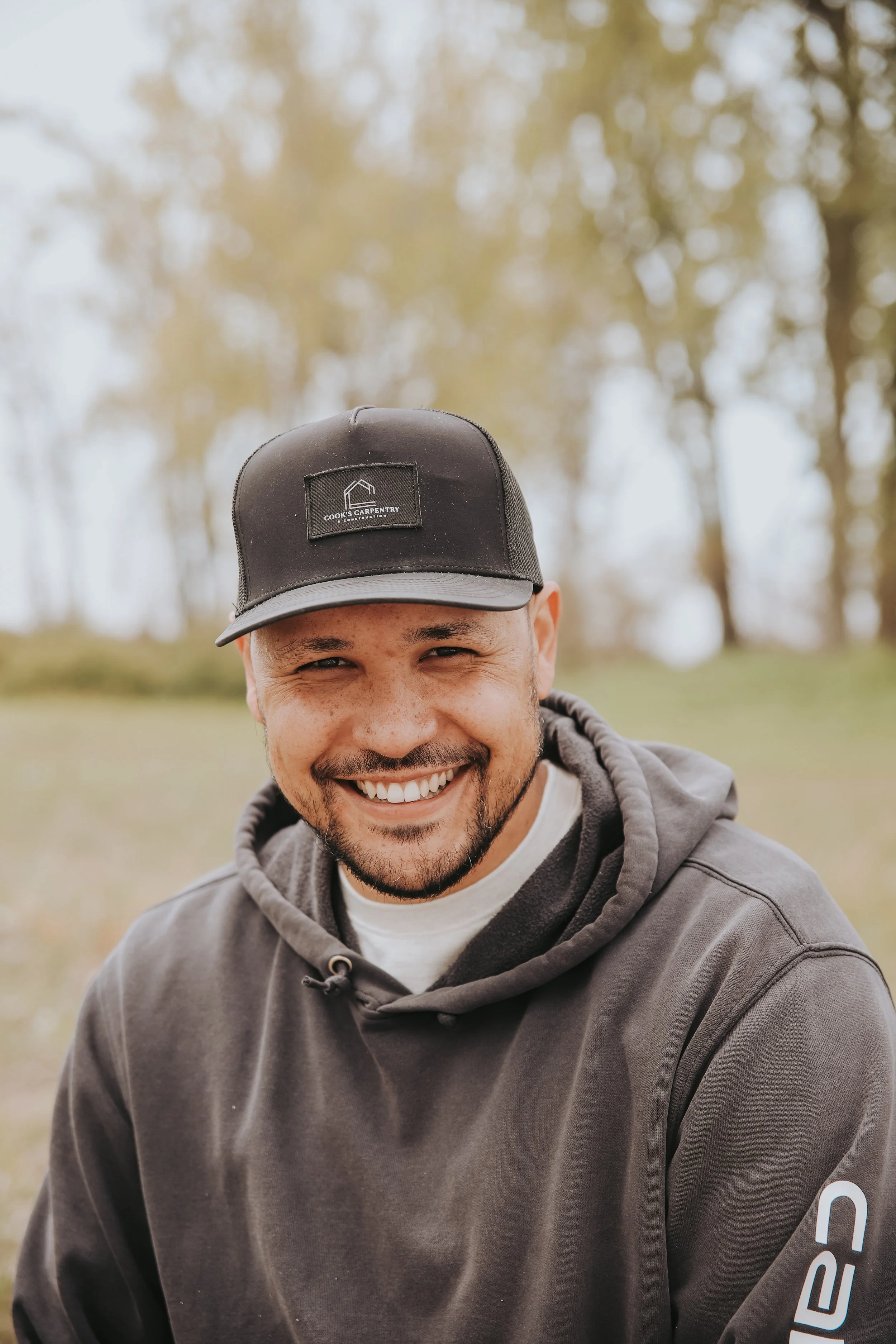 A smiling man with a beard and mustache wearing a black cap and a gray hoodie outdoors with blurred trees in the background.