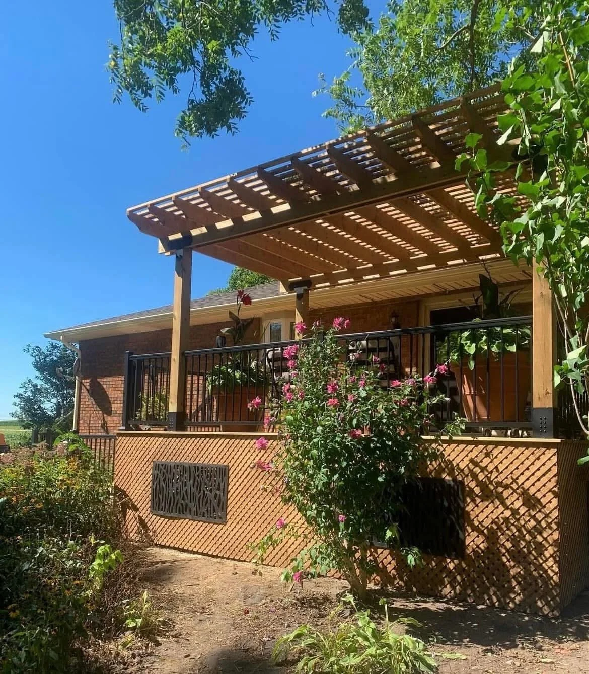 A house with a brick exterior and a wooden deck features a pergola-covered patio area adorned with pink flowering shrubbery and potted plants under a clear blue sky.