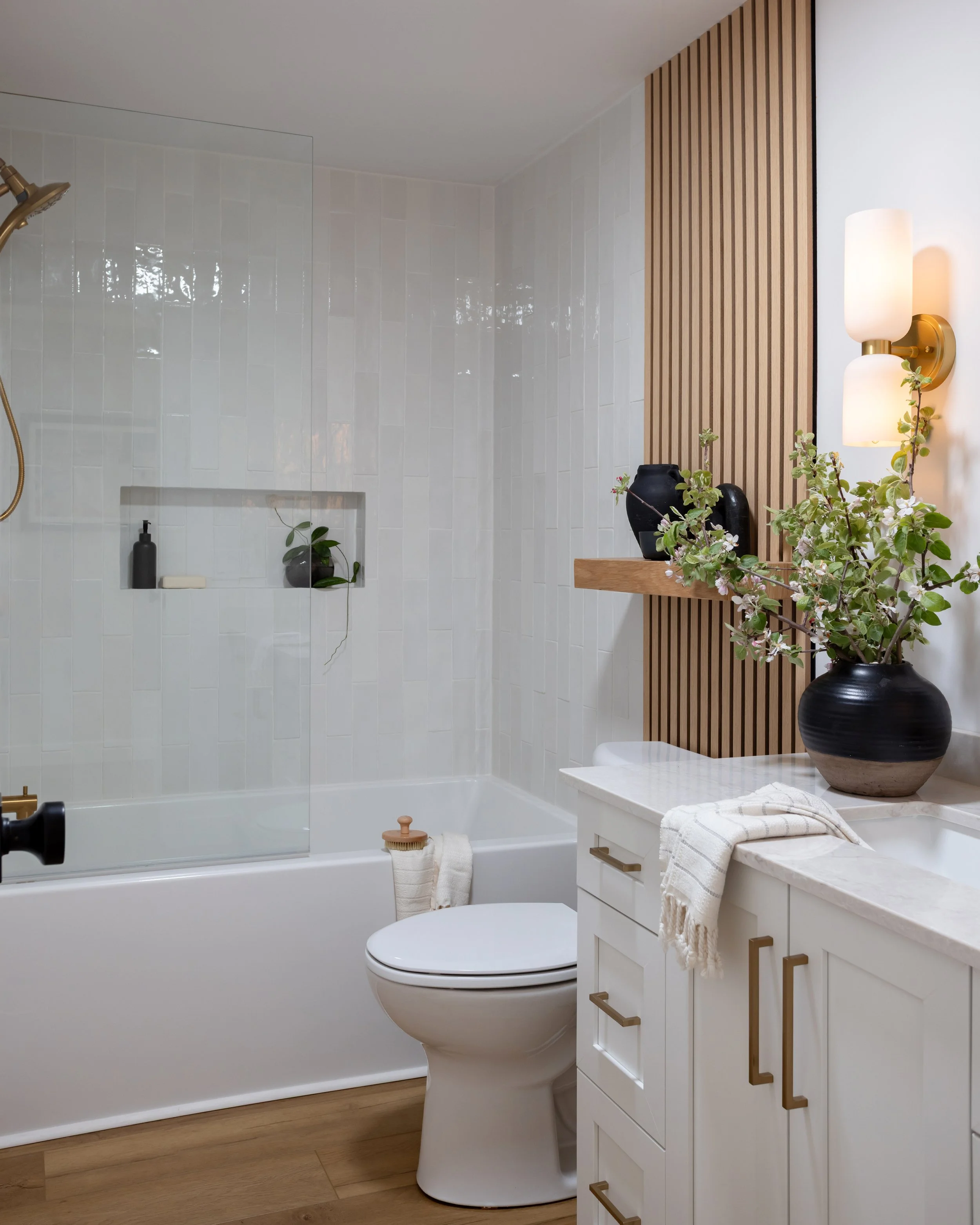 Modern bathroom with a shower area featuring white tiles, a built-in niche with a plant and soap, a wooden slat accent wall, decorative vases, a lamp, and a marble countertop with a towel.