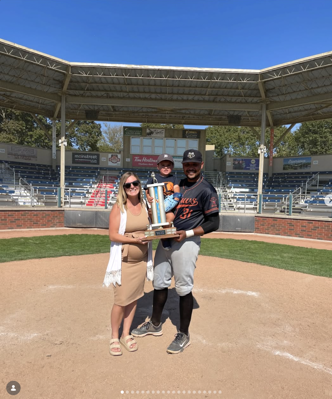 A woman, a young boy, and a baseball player are standing on a baseball field. The baseball player is holding a trophy, and all three are smiling at the camera. The stadium seating is visible in the background.