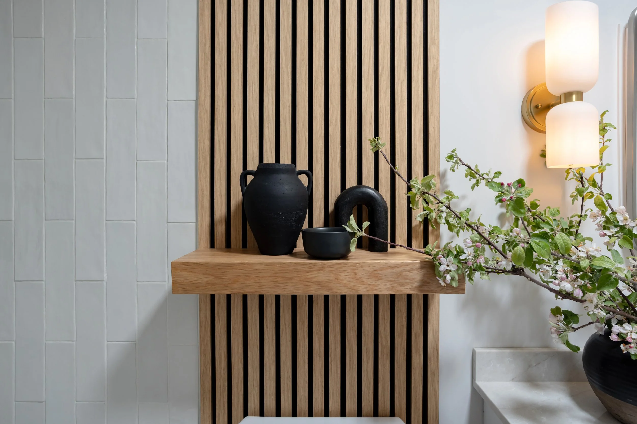 Decorative indoor wall with wooden slats, black vases, a branch with white flowers, a marble surface, and a modern wall lamp.