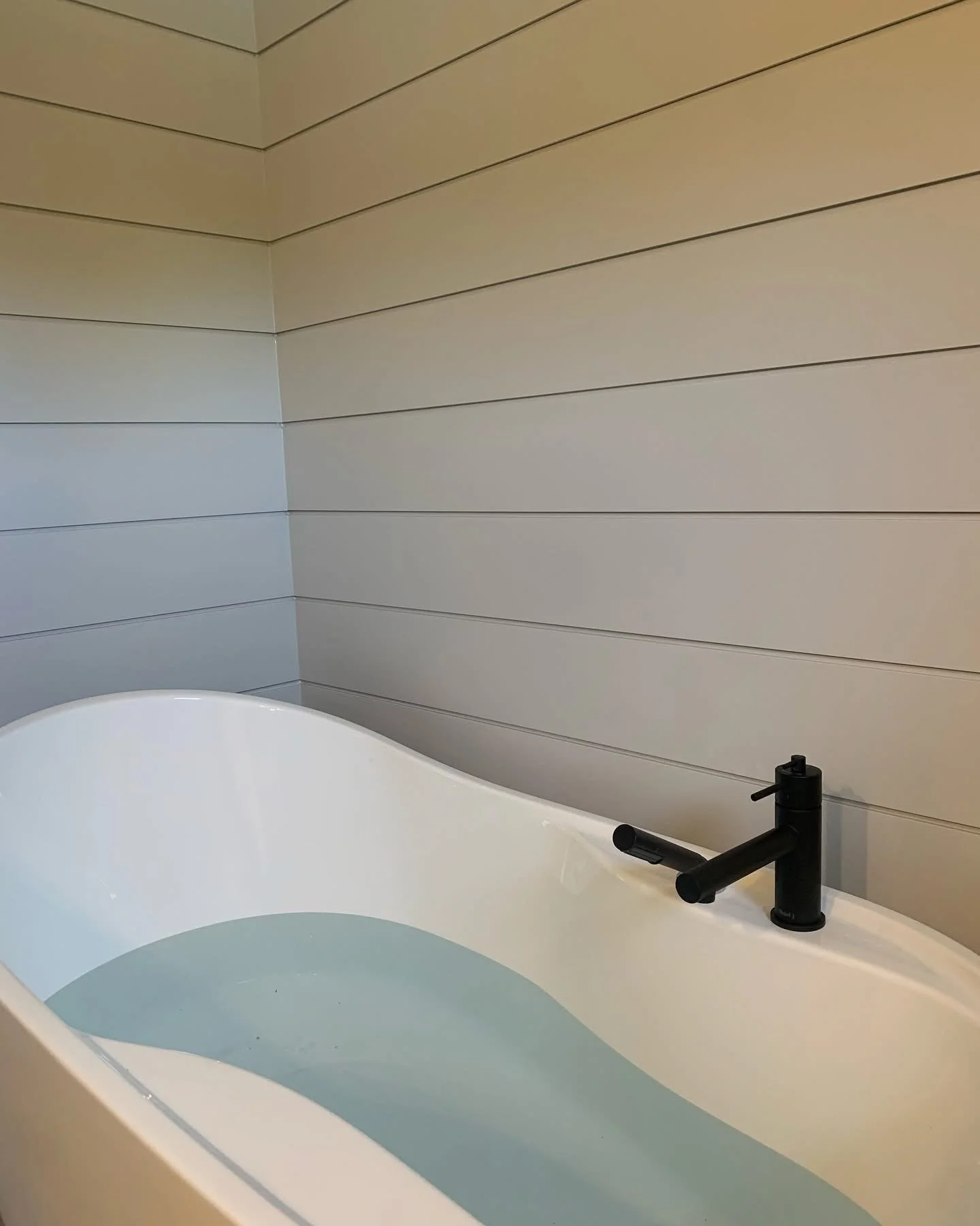 A white bathtub filled with water, set against a beige wall with horizontal shiplap paneling, and a black faucet with a single handle.