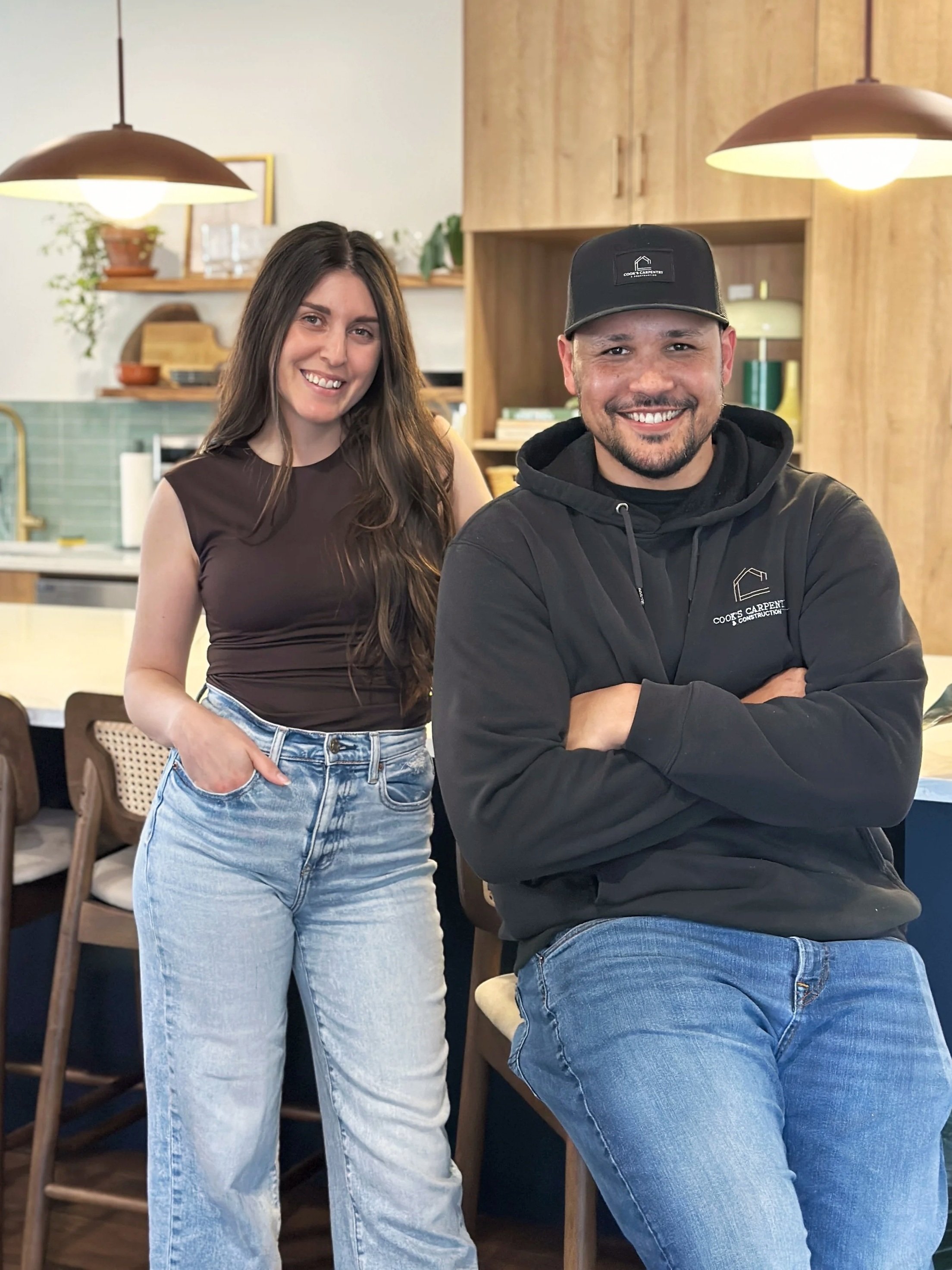A woman with long brown hair wearing a brown sleeveless top and light-wash jeans posing next to a man wearing a black hoodie and a black hat with a construction company logo, inside a modern kitchen with wooden cabinets and decorative shelves.