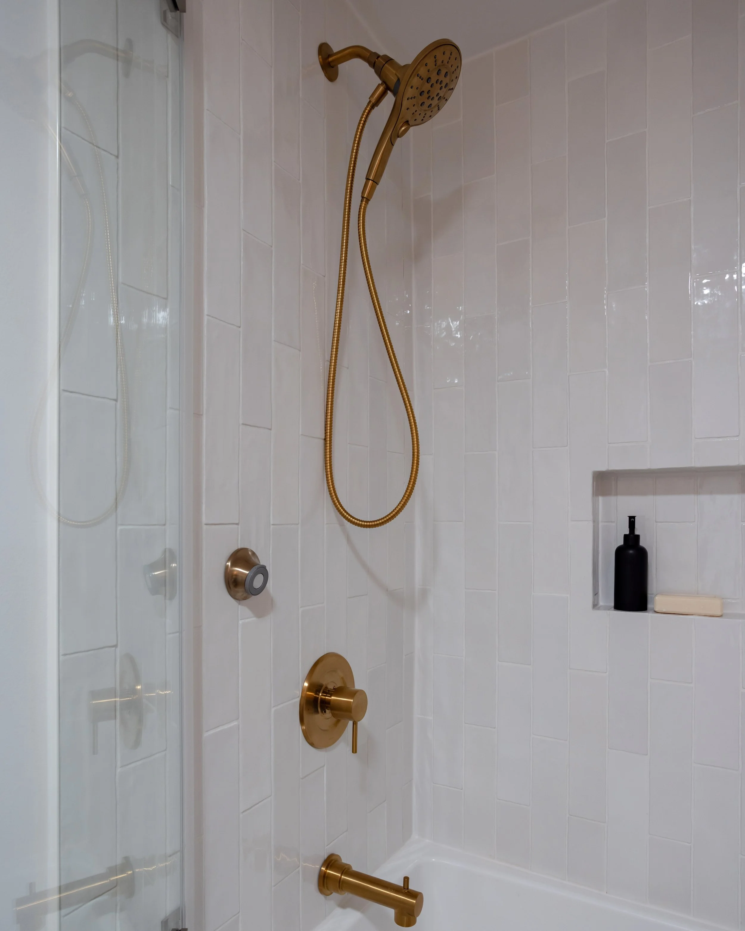 A shower area with a gold-colored showerhead, handheld attachment, faucet, and handle, all mounted on a white tiled wall. There is a black soap dispenser and a bar of soap on a built-in shelf.