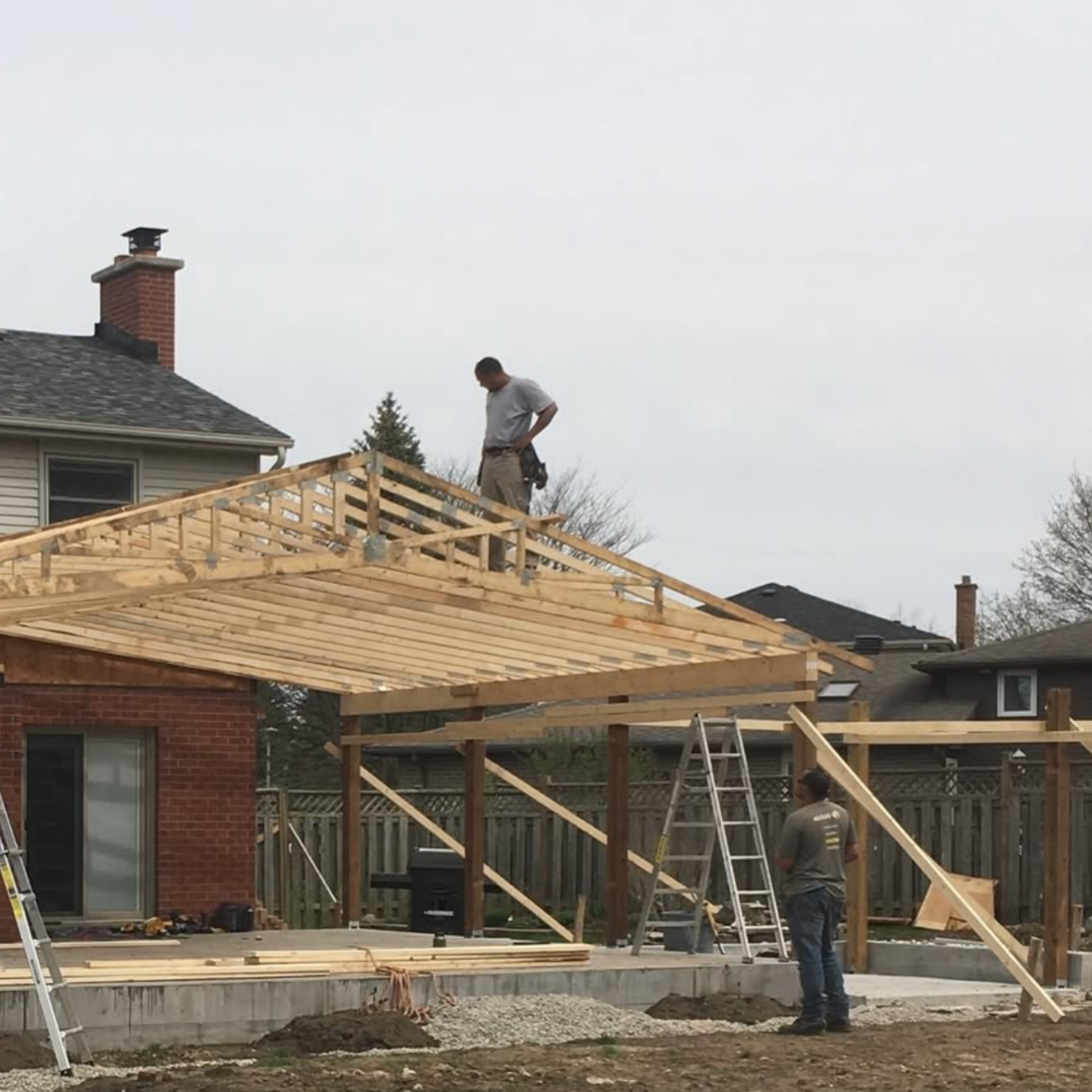 Construction workers building a wooden porch or deck extension on a house, with one worker on the roof and another on the ground near a ladder, during daytime.