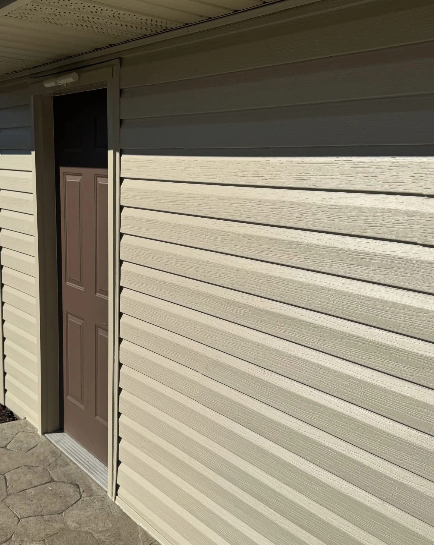 Exterior view of a house with beige vinyl siding, a brown front door, and a small outdoor light fixture at the top left of the door frame.
