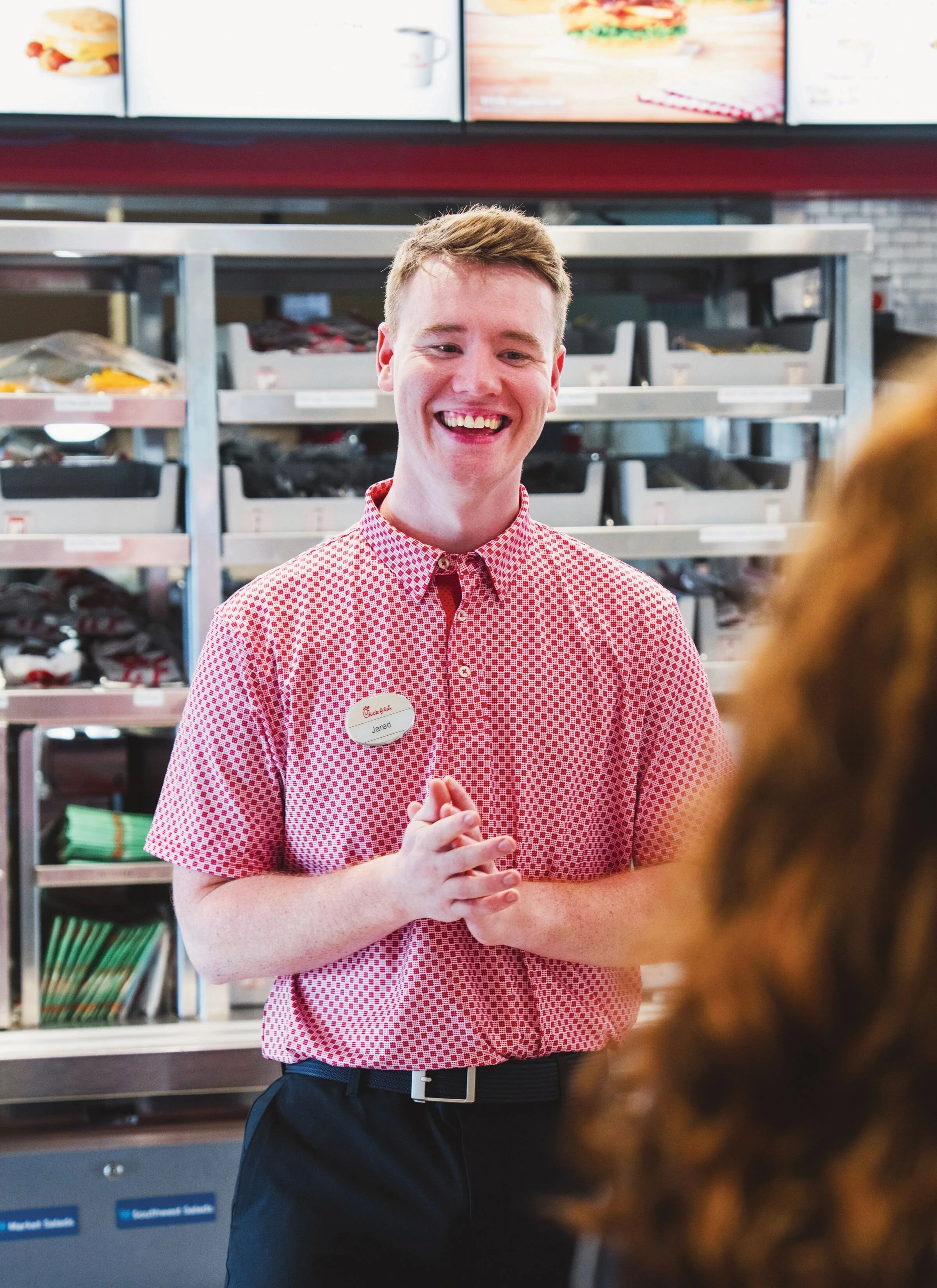 A young man wearing a red and white checkered shirt with a name tag that reads Jared, smiling and clasping his hands together, standing in front of a display shelf with snacks and food items in a store.