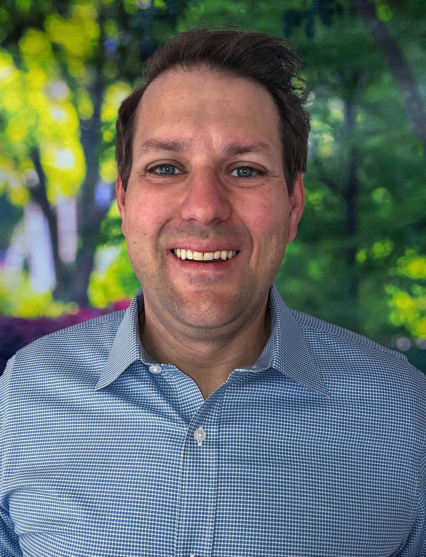 Close-up of a smiling man with blue eyes wearing a checkered blue shirt, standing in front of a blurred background of green trees and foliage.