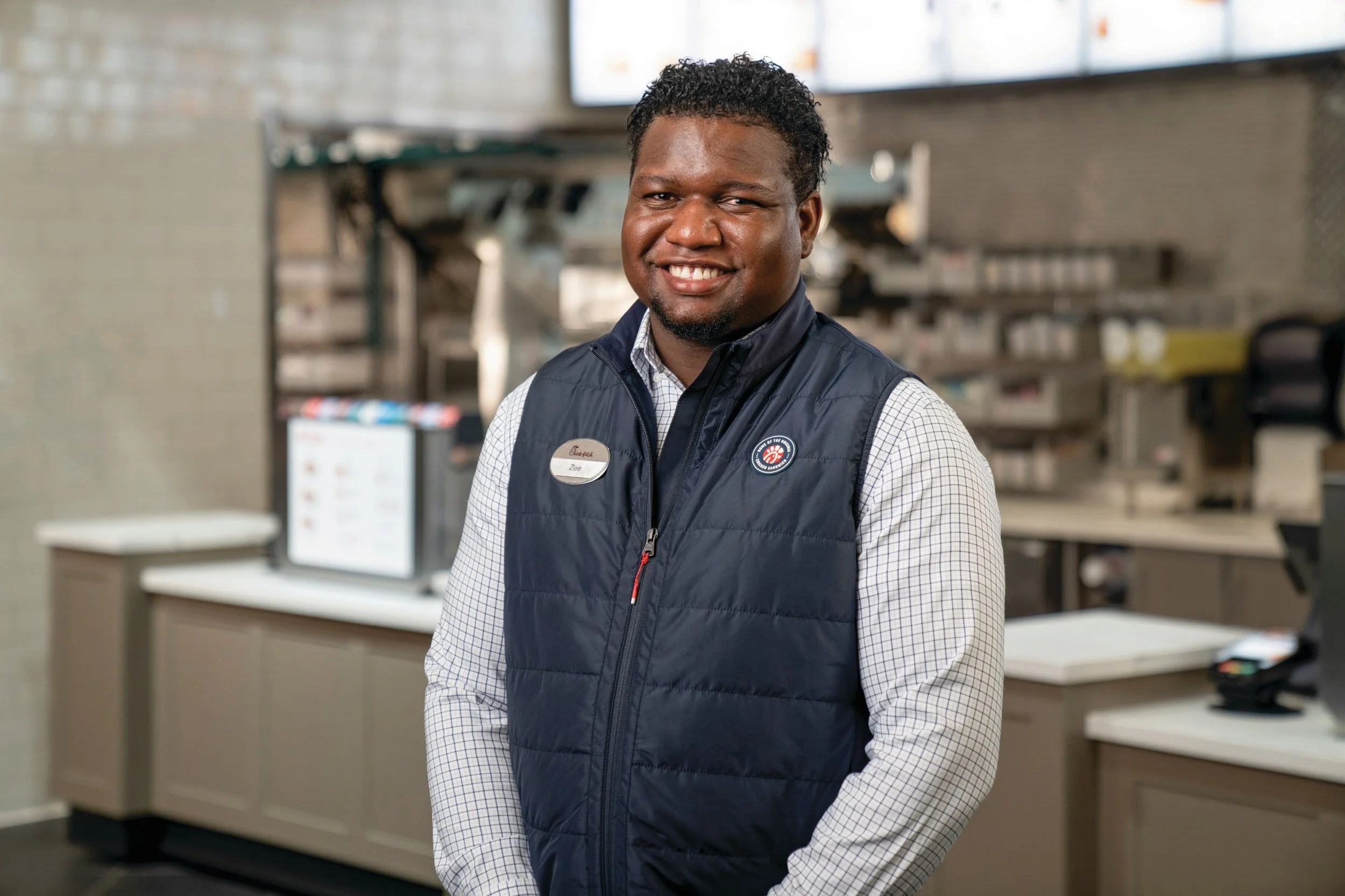 A man with curly black hair smiling, wearing a navy vest with a Chick-fil-A name tag, standing inside a fast-food restaurant.