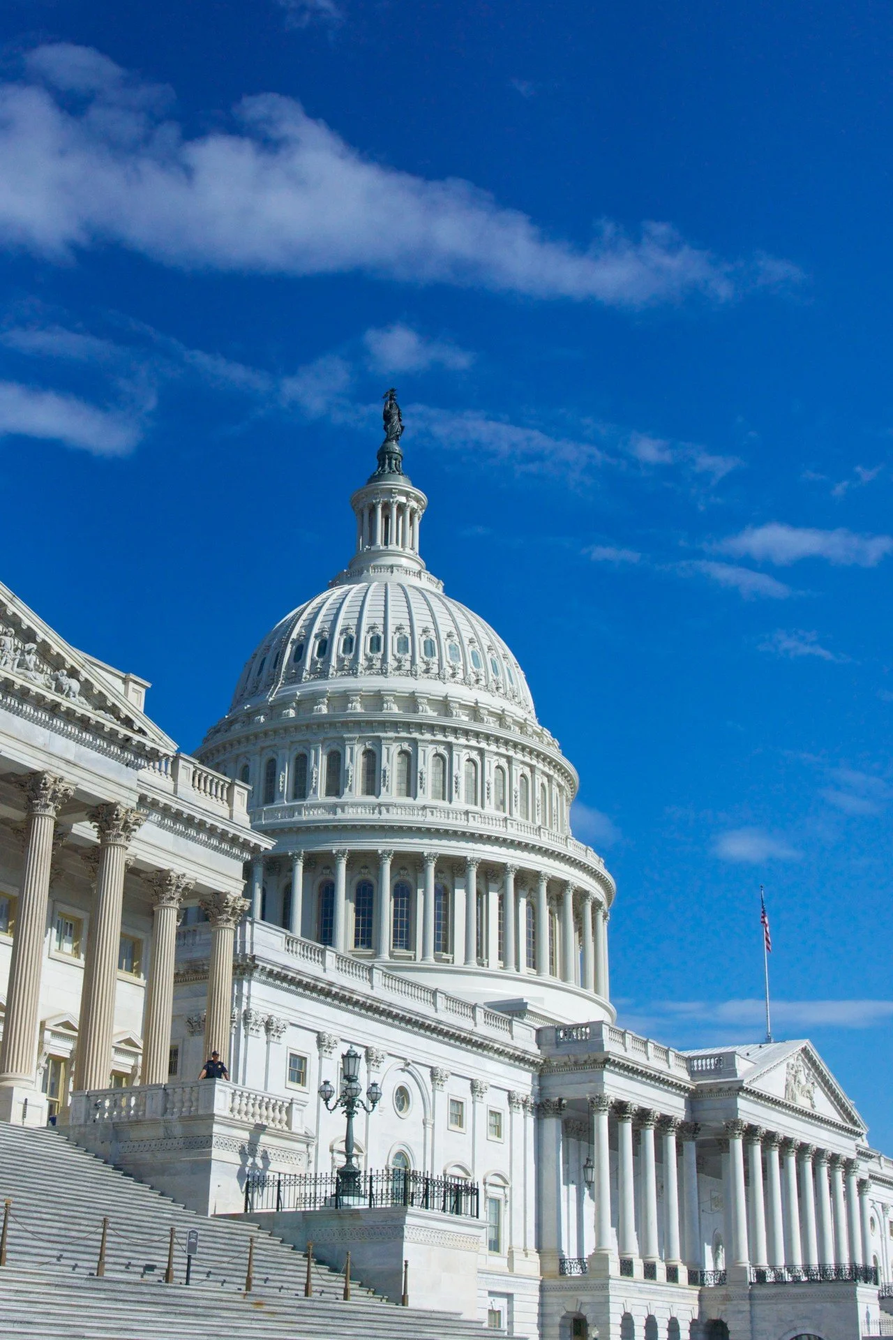 The United States Capitol building with its large white dome and a flagpole with the American flag, under a blue sky with some clouds.