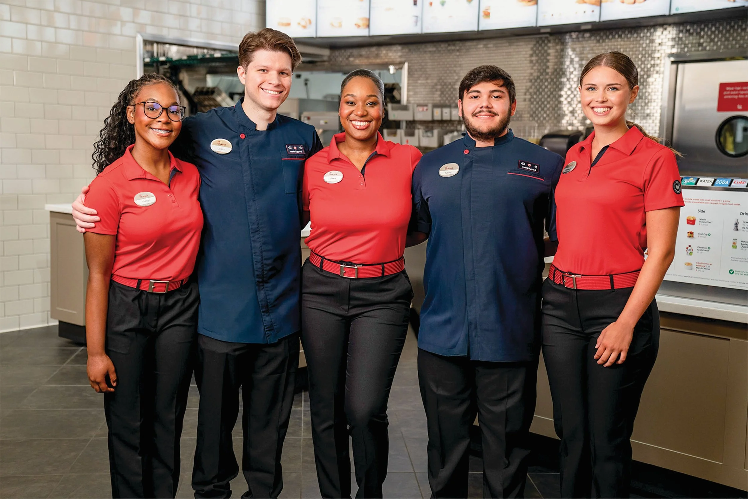 Group of five diverse employees standing together inside a fast food restaurant, smiling, wearing red and navy uniforms with name tags.