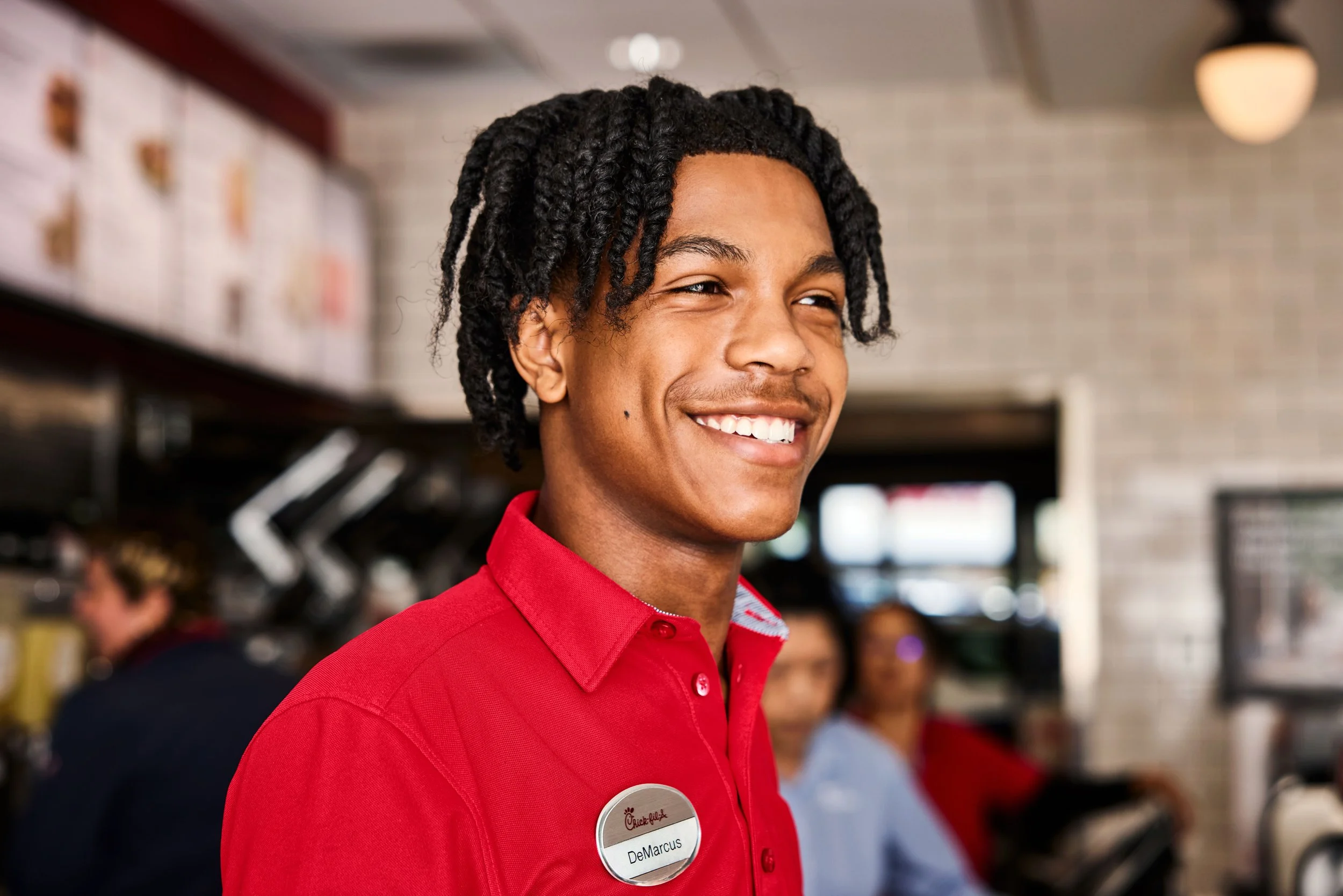 A young man wearing a red Chick-fil-A uniform with a name tag smiling in a busy restaurant.