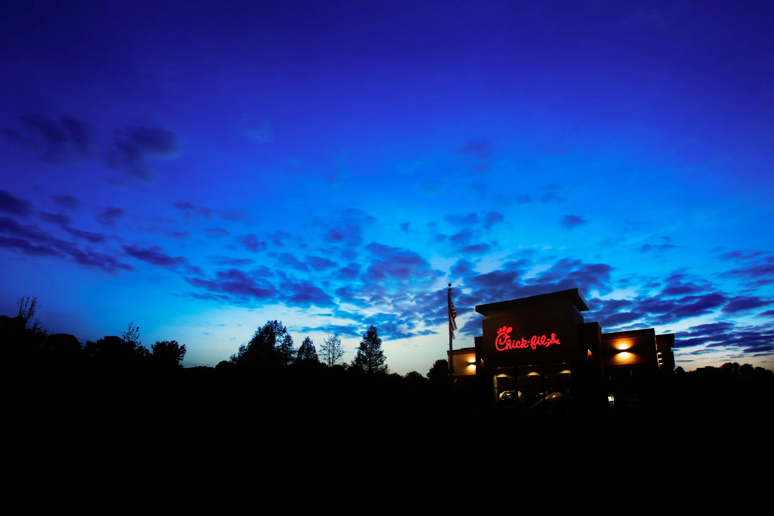 Sky with scattered clouds at dusk, silhouette of trees and a Chick-fil-A restaurant with illuminated sign and flag in foreground.