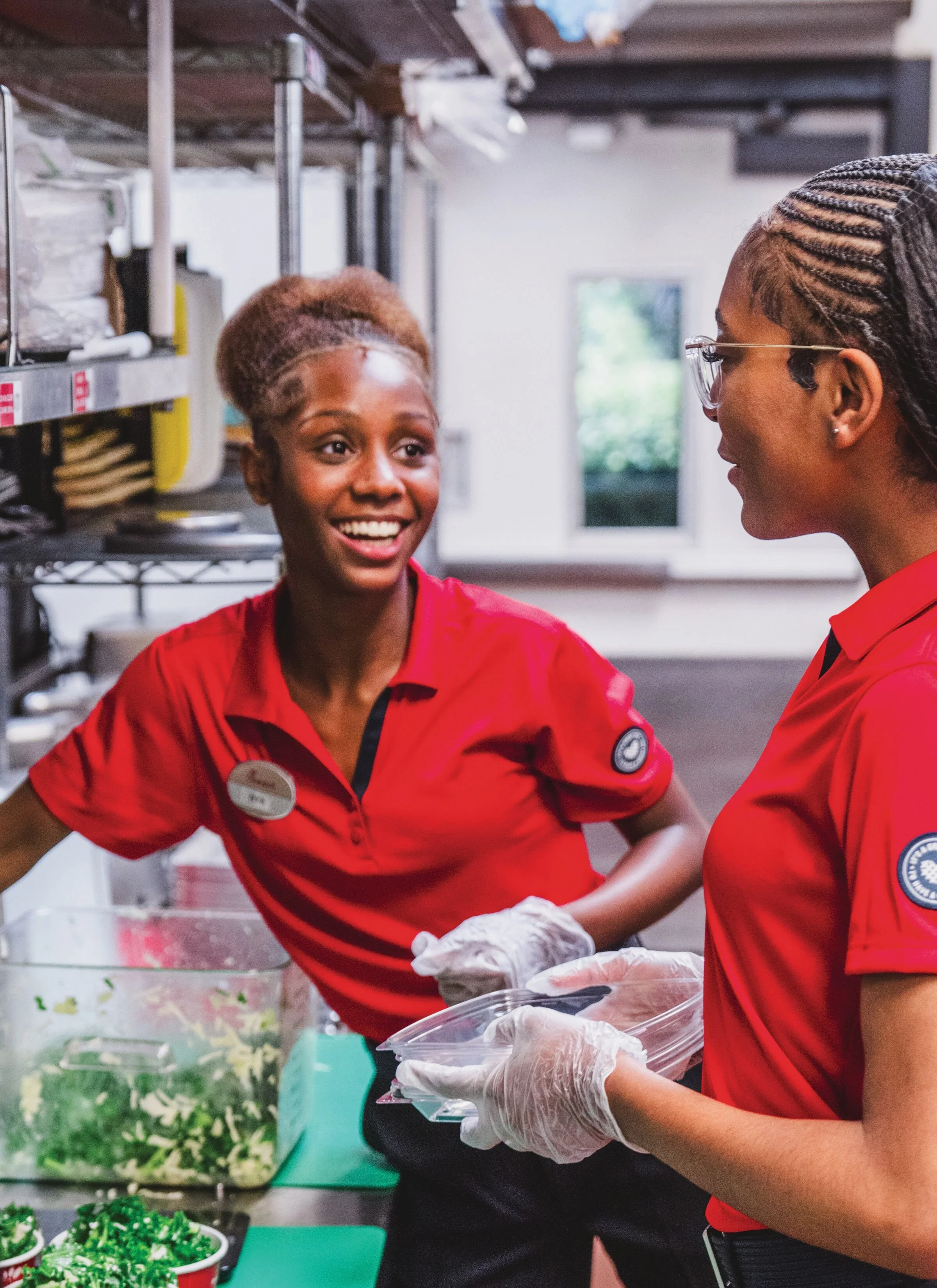 Two women in red uniforms preparing food in a kitchen, with vegetables on a green cutting board and containers, wearing gloves and chef badges.