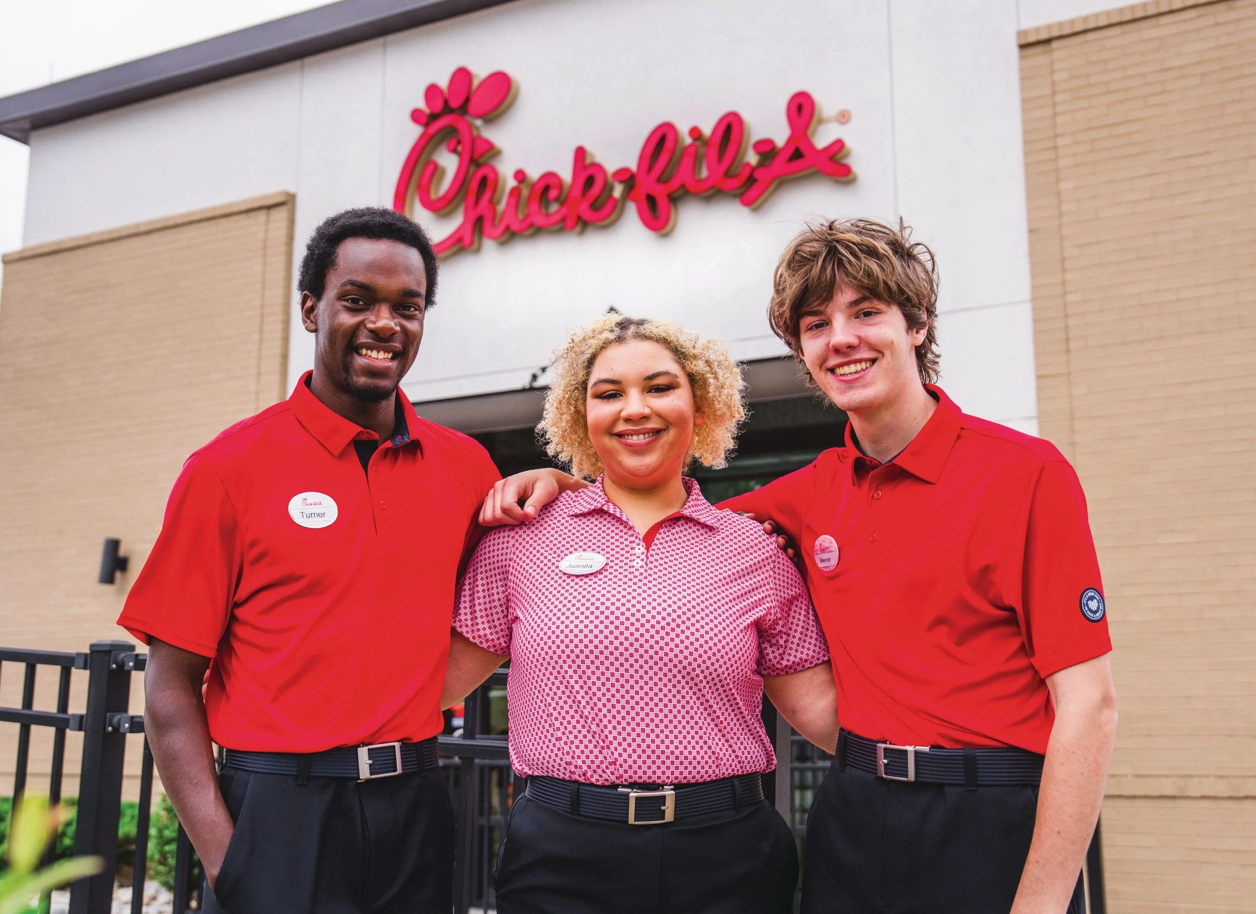 Three Chick-fil-A employees standing outside by a black railing in front of the restaurant, smiling. The employees are wearing red Chick-fil-A uniforms with name tags. The building has a large Chick-fil-A logo on the wall behind them.