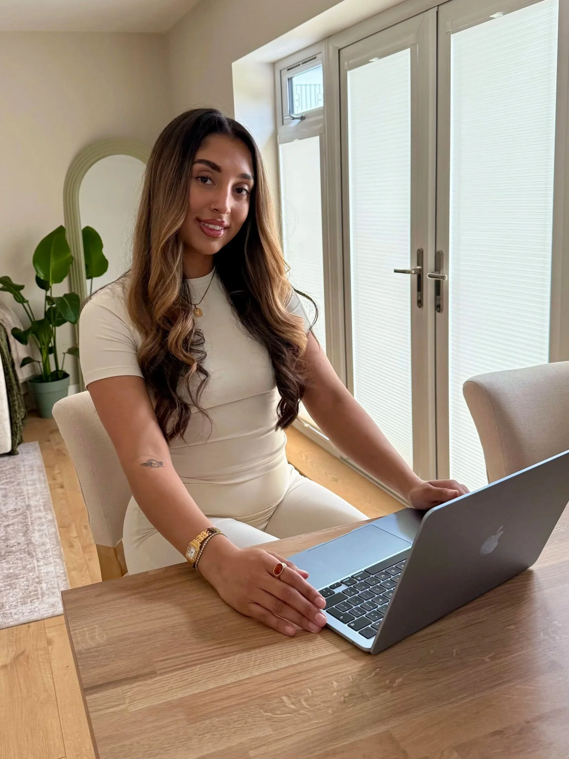 A woman with long wavy hair, wearing a white dress and jewelry, sitting at a wooden table with a silver laptop in front of her in a bright room with large glass doors and a plant in the background.