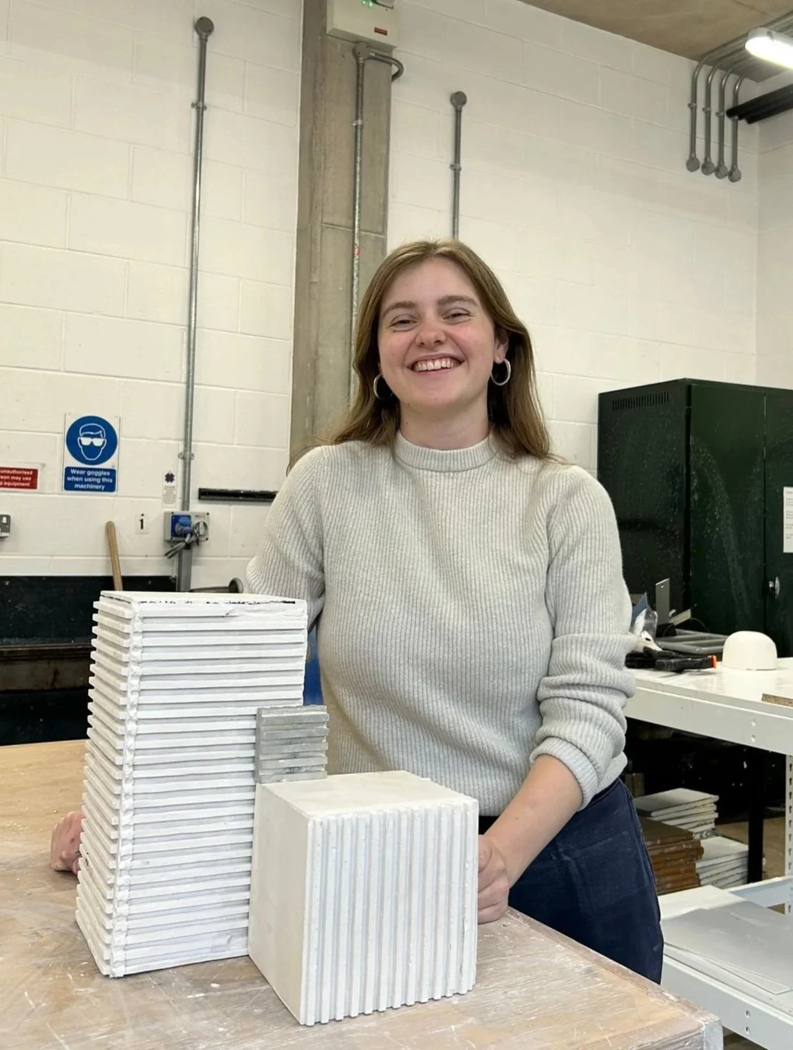 A woman with brown hair, wearing a light gray sweater and hoop earrings, standing in a workshop surrounded by ceramic tiles. She is smiling, with several stacks of tiles on a wooden table in front of her, and industrial equipment visible in the background.