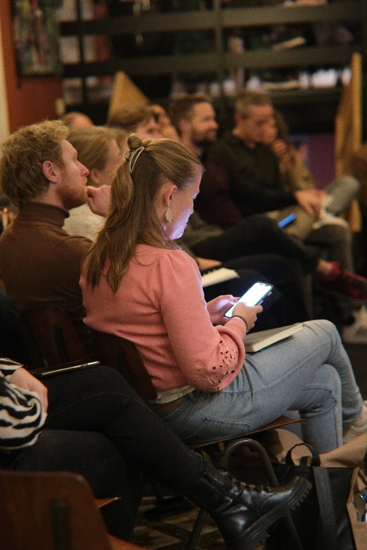 Mensen zitten op stoelen in een zaal, de vrouw in de voorgrond kijkt op haar telefoon.
