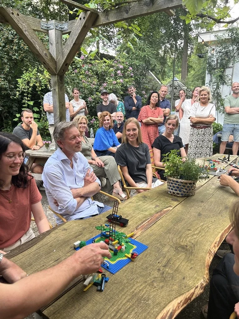 Een groep mensen zit en staat buiten in een tuin, terwijl ze naar iemand kijken die LEGO-bouwwerken op een houten tafel bouwt. Er is groene planten en bloemen op de achtergrond.