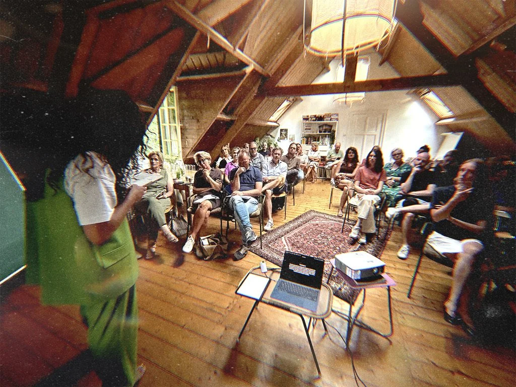 Een groep mensen zit in een knusse, zolderkamerachtige ruimte met houten muren en plafond. Ze luisteren naar een spreker die op een scherm bezig is met een presentatie. Er liggen boeken en een projector op een tafel vooraan.