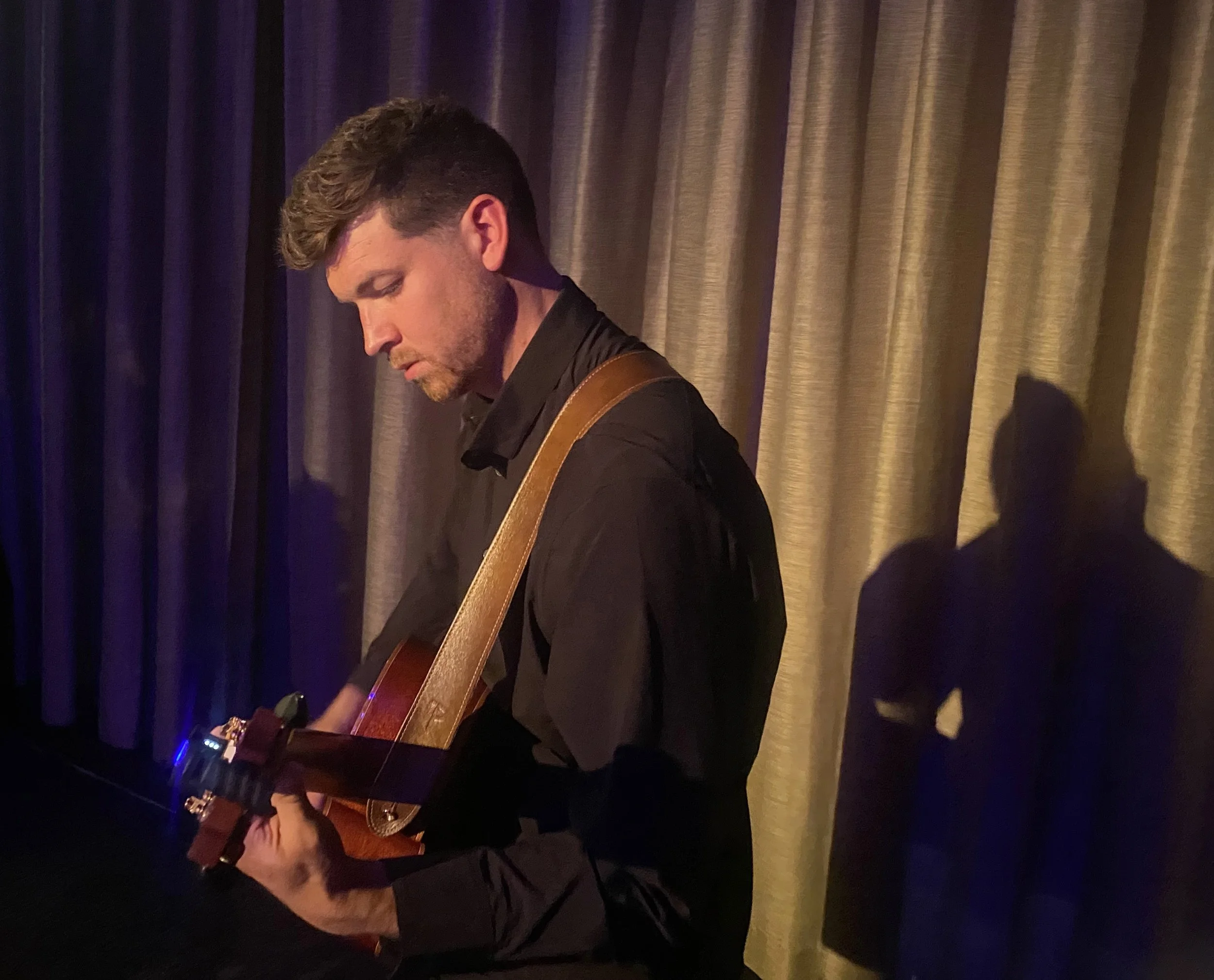 A man playing an acoustic guitar in front of a backdrop of dark purple and gold curtains.