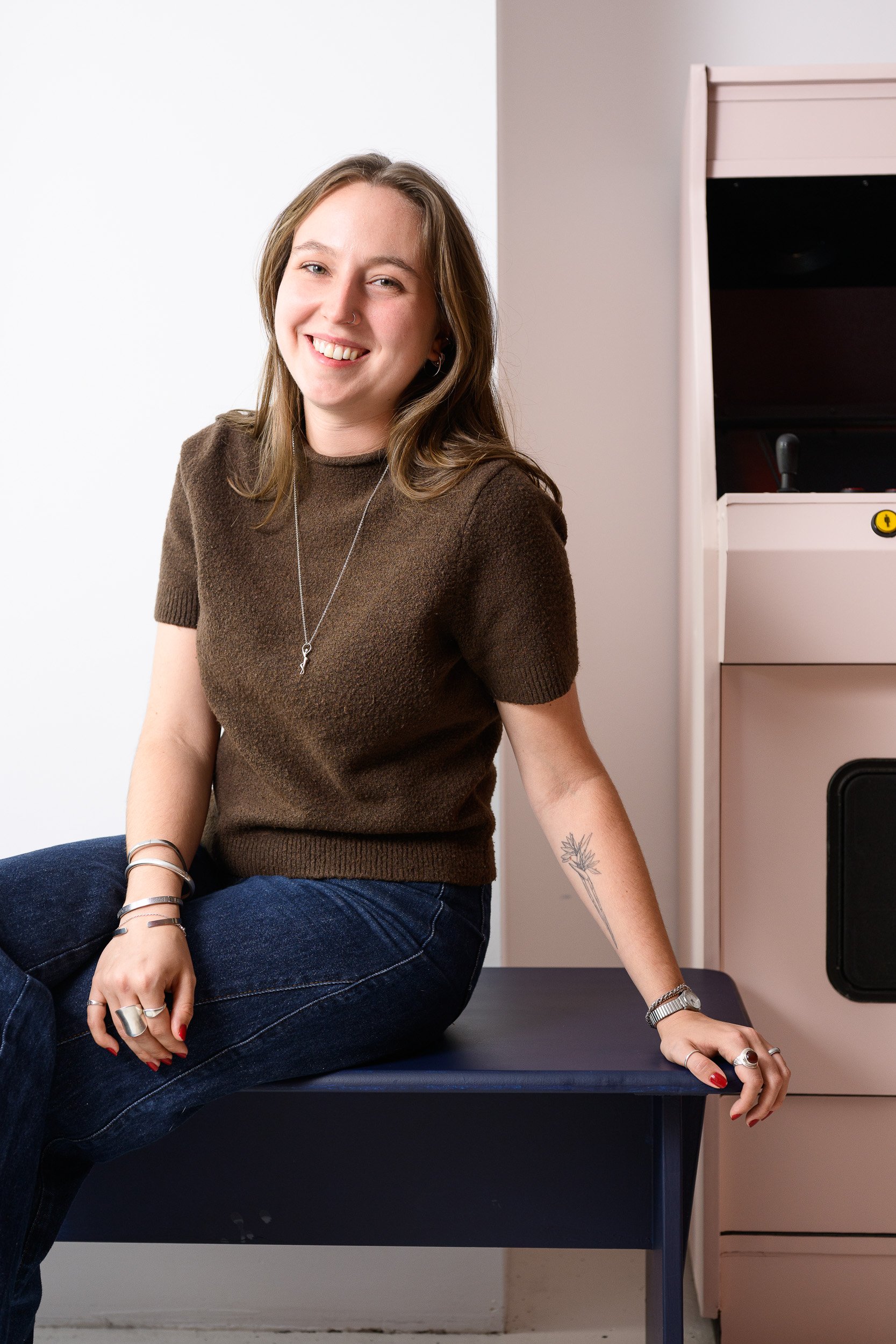 een jonge vrouw met lange bruin haar, zittend op een tafel, lachend naar de camera, gekleed in een bruin trui en donkerblauwe jeans, met sieraden en een tattoo op haar linker onderarm, naast een rode jukebox in een interieur.