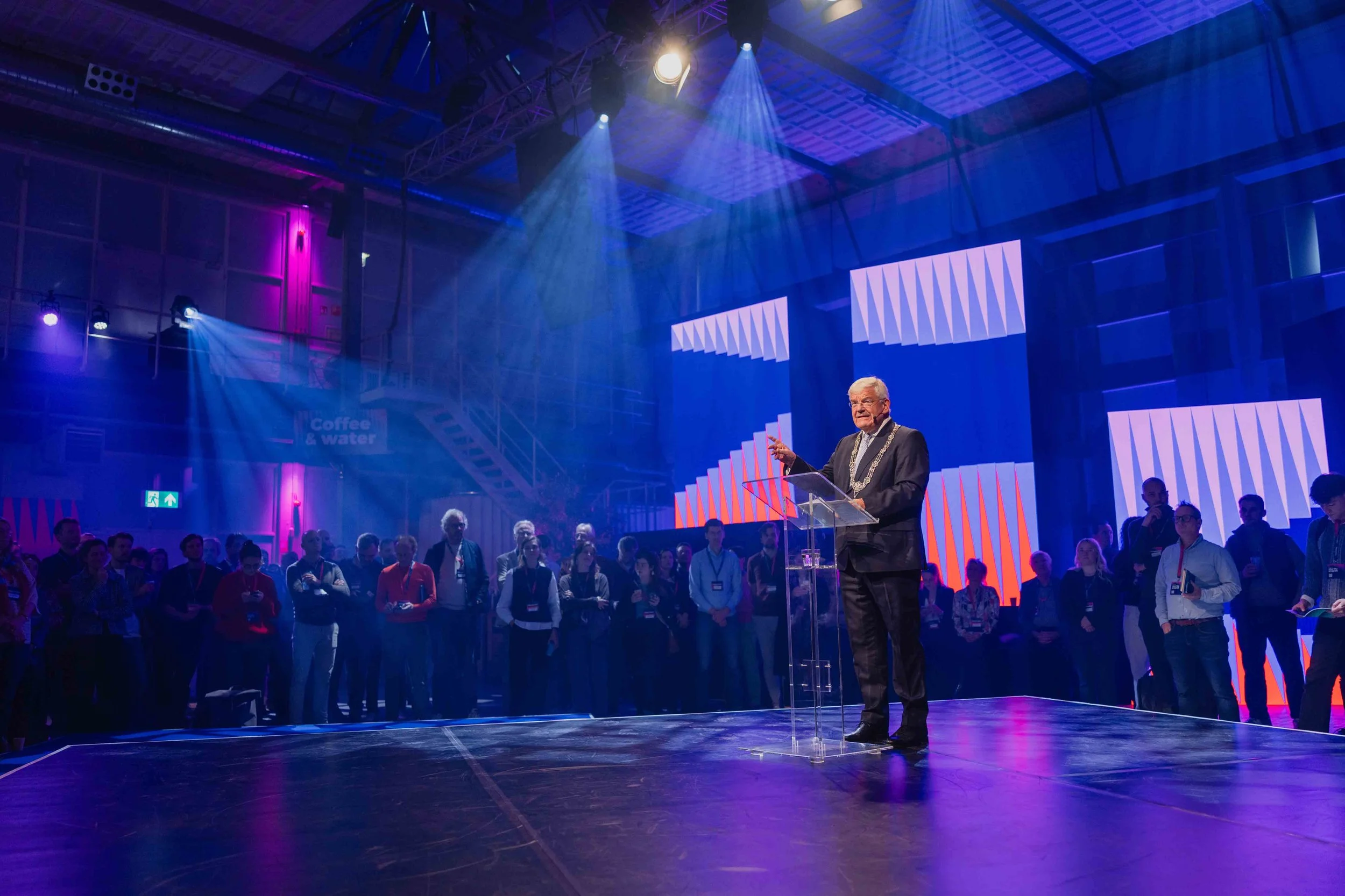 Een man in een donker pak met een scherm op de achtergrond staat aan een spreekgestoelte tijdens een evenement. Er is een groep mensen die geluisterd en gekeken naar de spreker. De ruimte is verlicht met blauwe en paarse lichten, en er zijn grote schermen op de achtergrond.