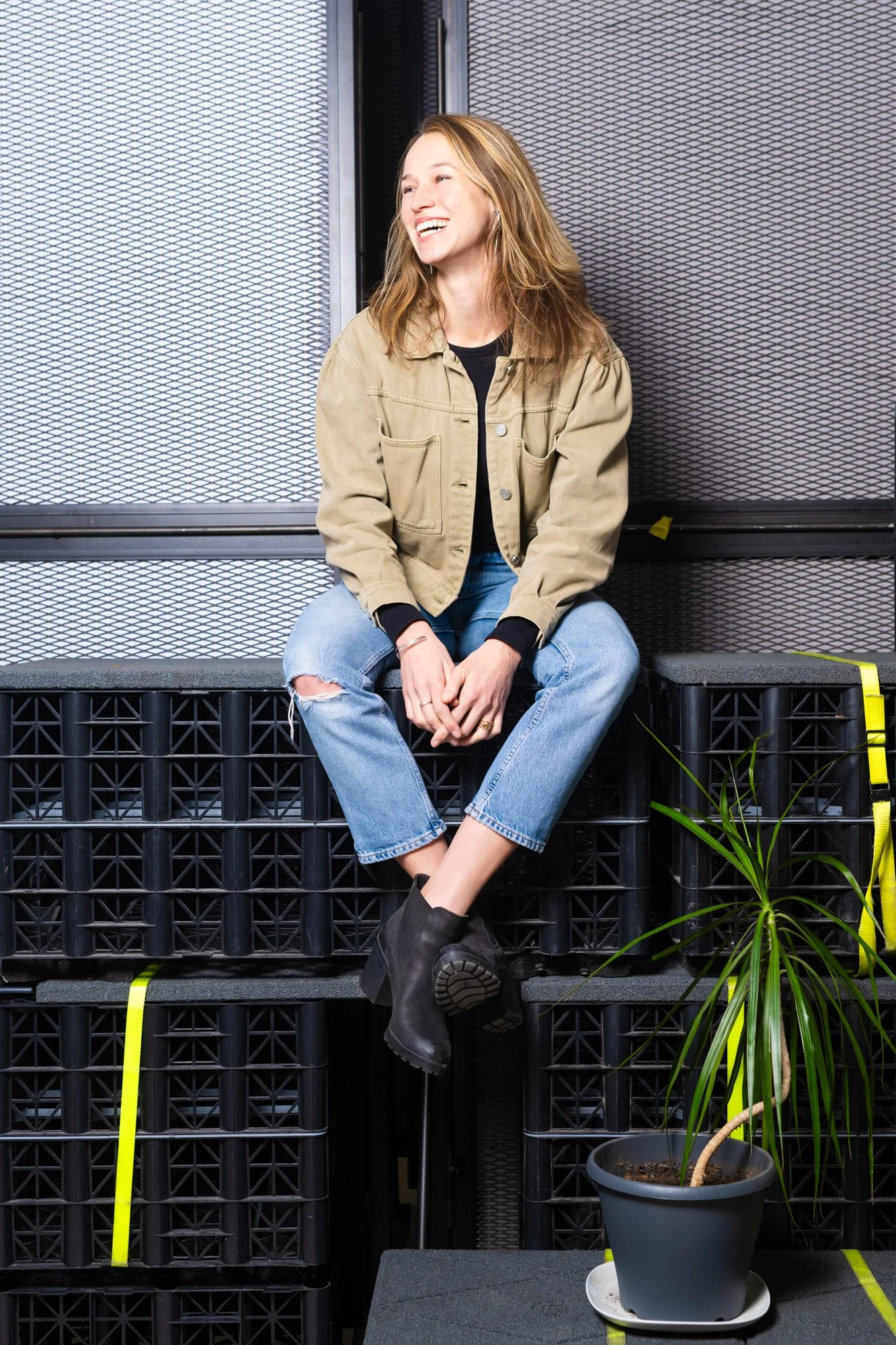 Een lachende vrouw met bruin haar, gekleed in een beige jas en blauwe jeans, zit op een stapel zwarte plastic kratjes in een ruimte met metalen achtergrond en een plant in een zwarte pot.