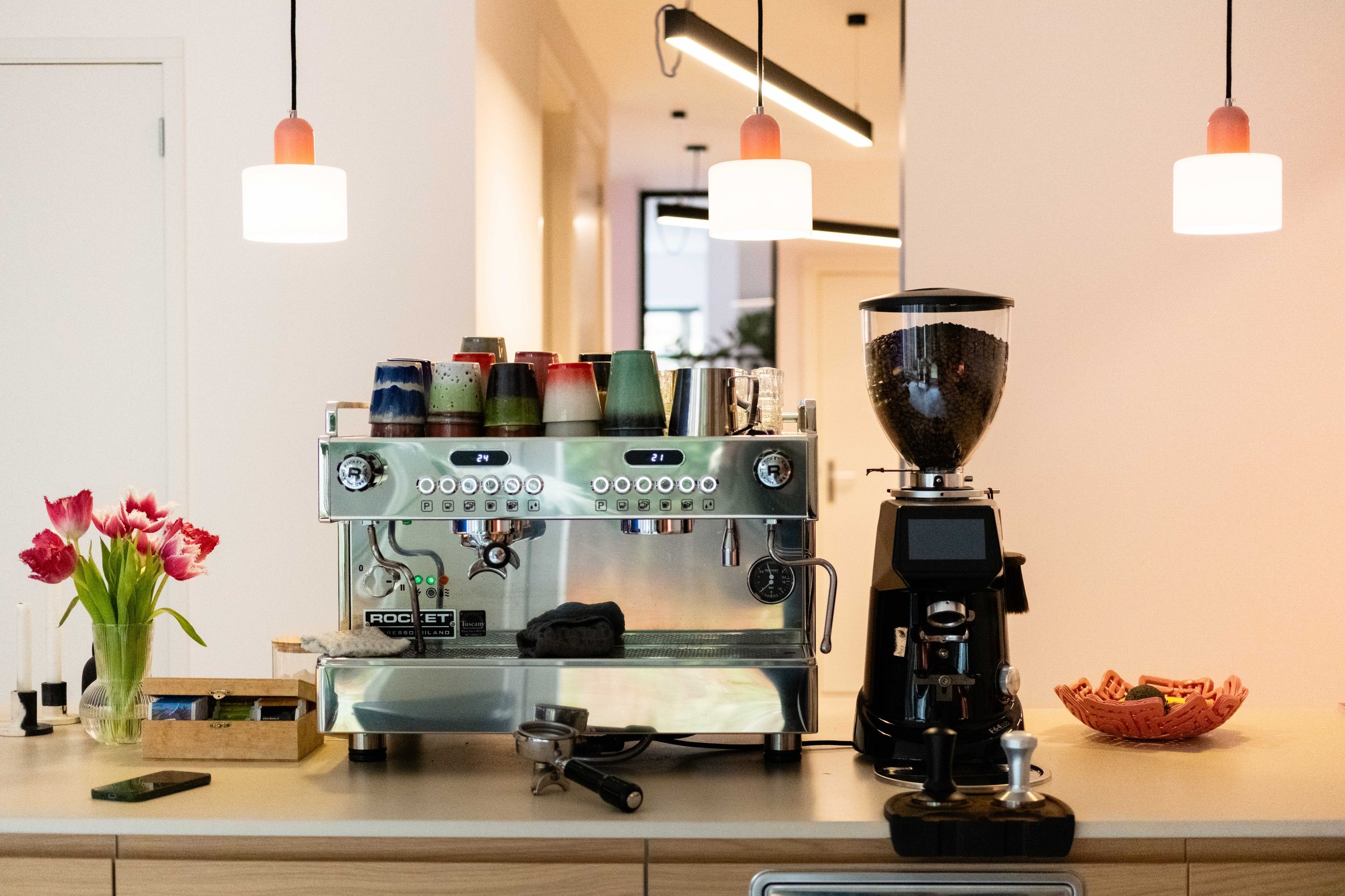Een moderne keuken met een koffiezetapparaat, een kopje, een mes, een bord en een vaas met roze tulpen op een aanrecht. In de achtergrond hangen hanglampen en een keukenblok.