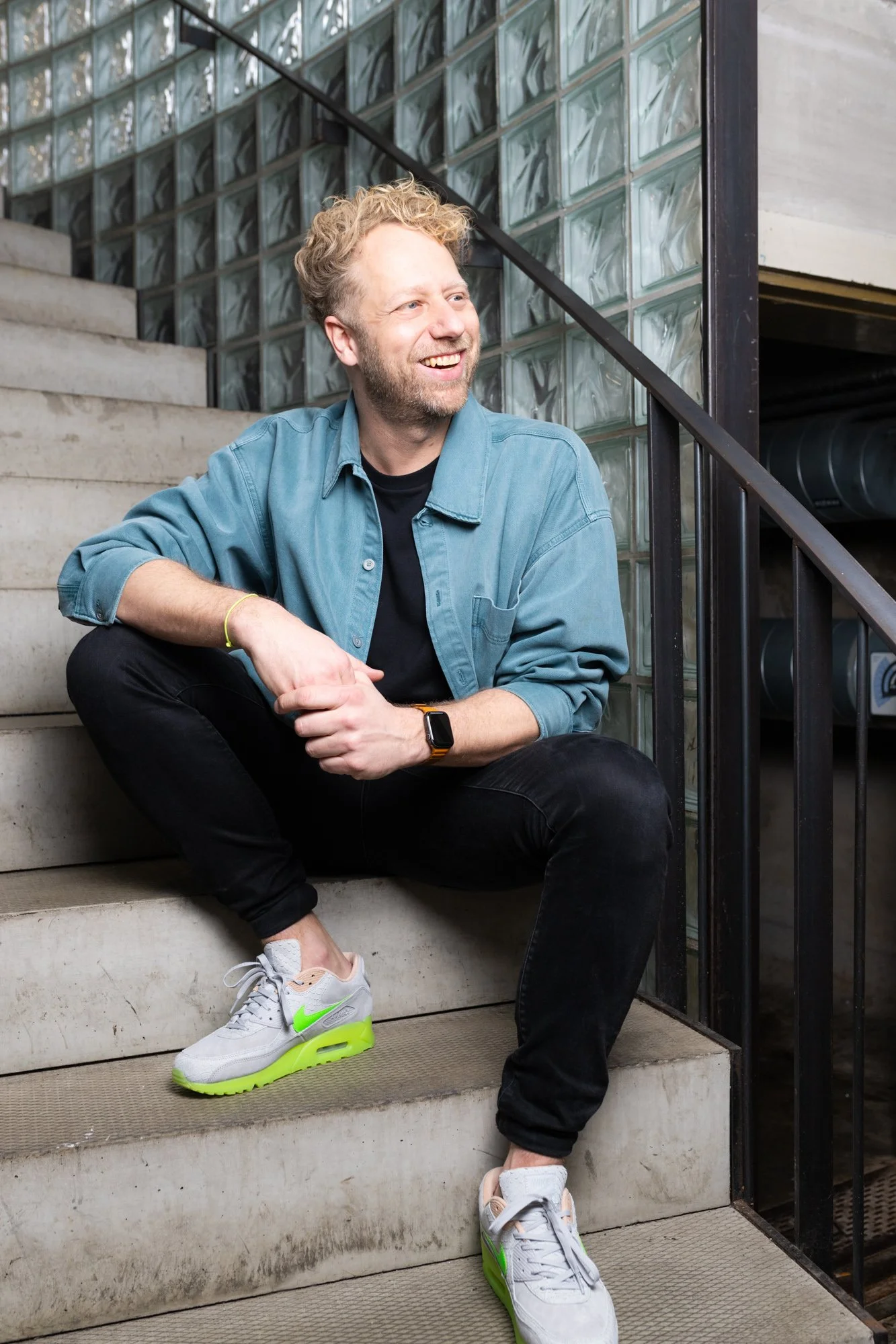 Man zit op de trappen buiten, lachend, met een denim jas en sneakers. Achter hem glasblokken en een metalen leuning.