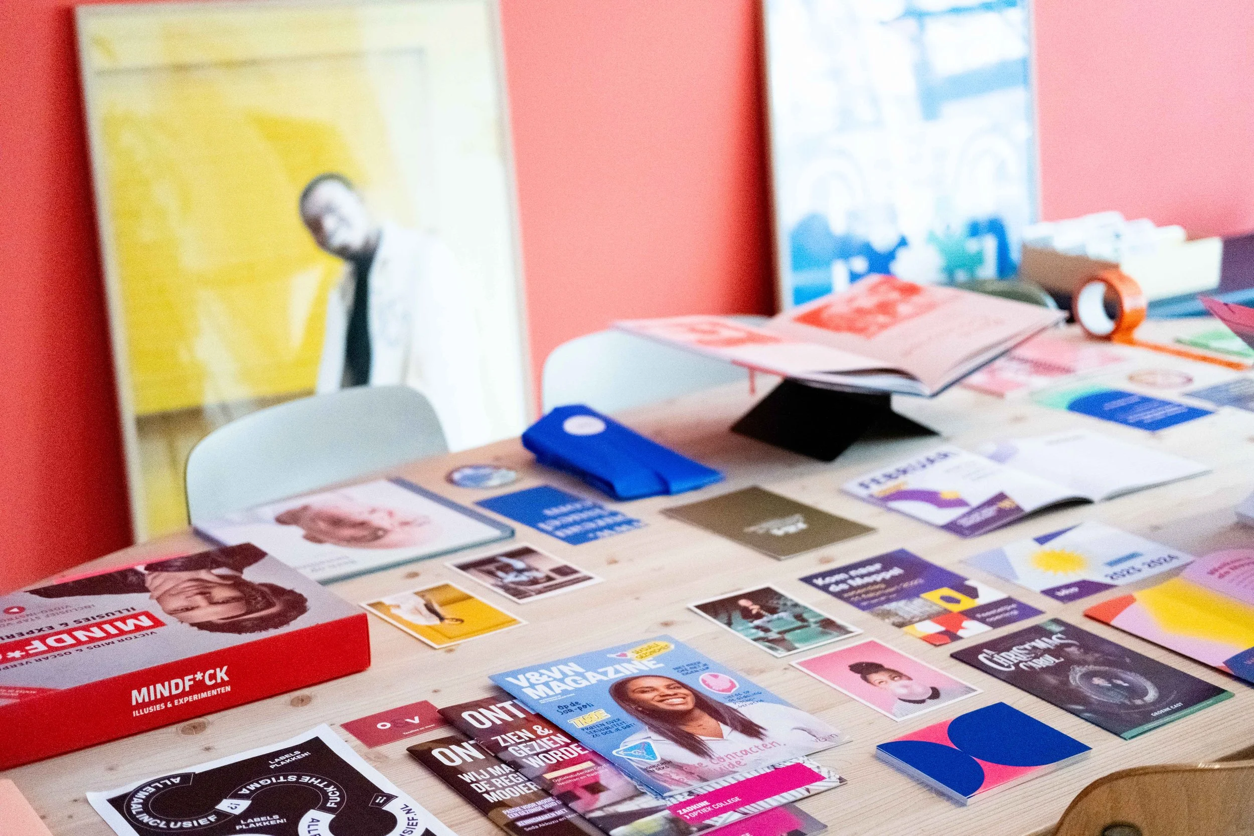 Een tafel met diverse flyers en brochures, met een rode doos op de voorgrond. Op de achtergrond zijn posters en een raam zichtbaar in een kleurrijke ruimte.
