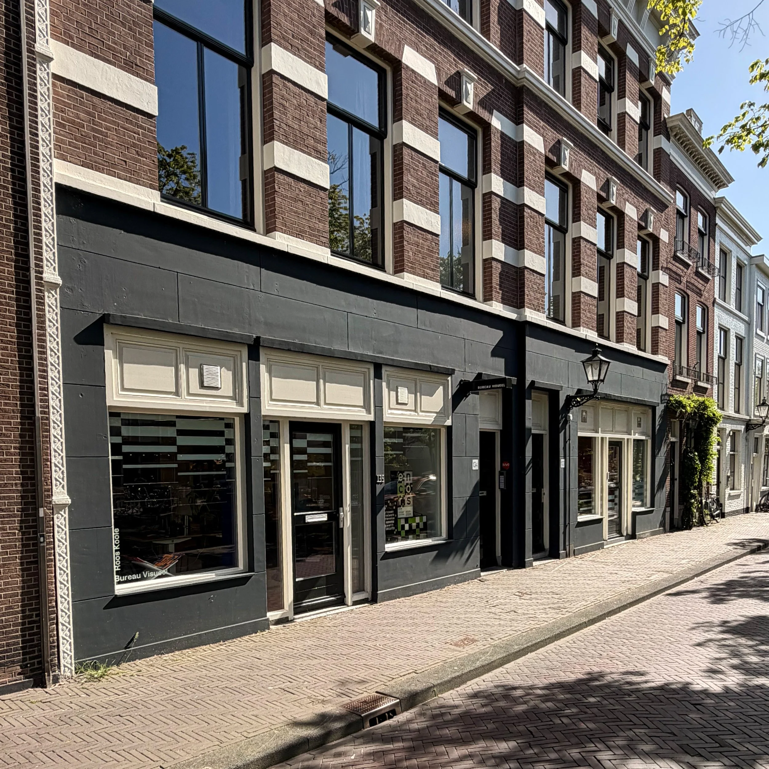 Een gevel van een geelbakstenen gebouw met grote ramen en een deur onder een zwart dak, gelegen aan een straat met een betegelde voetgangerszone en een lantaarnpaal.