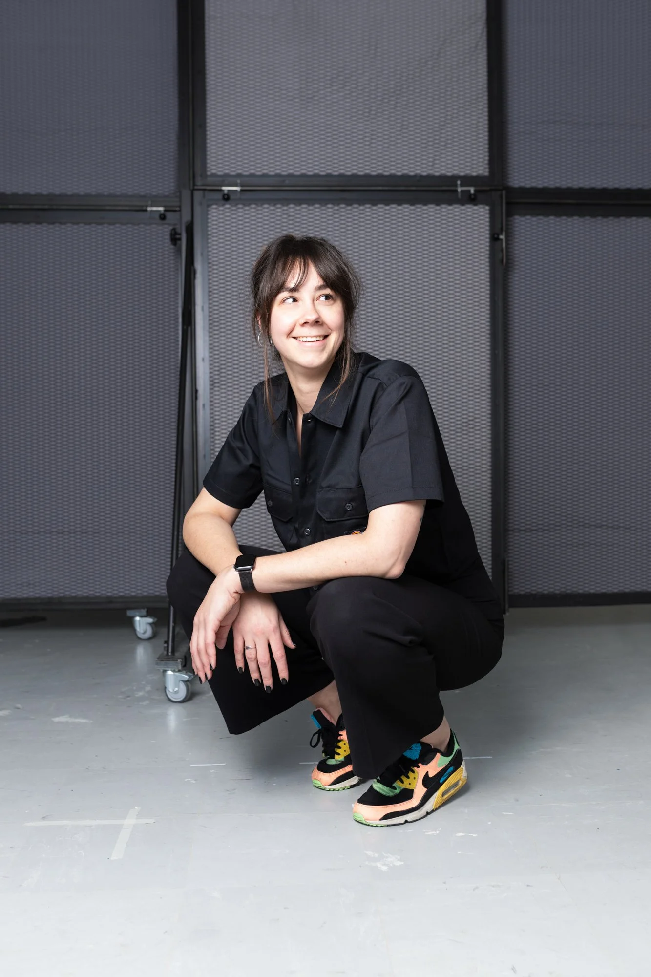 Vrouw in zwart overhemd en sneakers die in een halletje met metalen panelen en wieltjes zit.