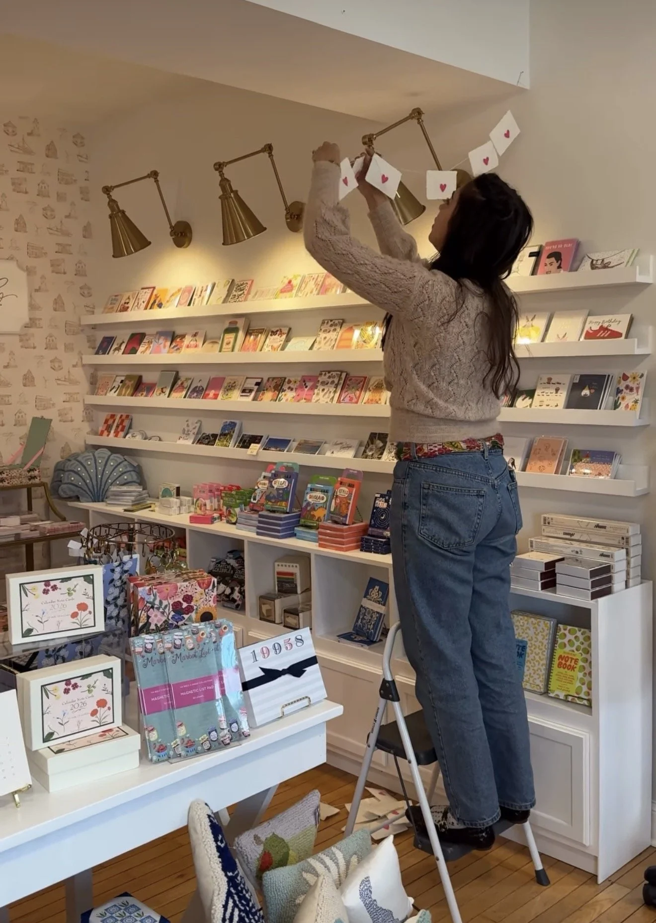 A woman with long dark hair, wearing a beige sweater and blue jeans, stands on a small step ladder arranging paper cards with red hearts on a ceiling hook in a cozy bookstore. The store has colorful books and stationery on white shelves and tables, with decorative pillows and framed artwork on display.