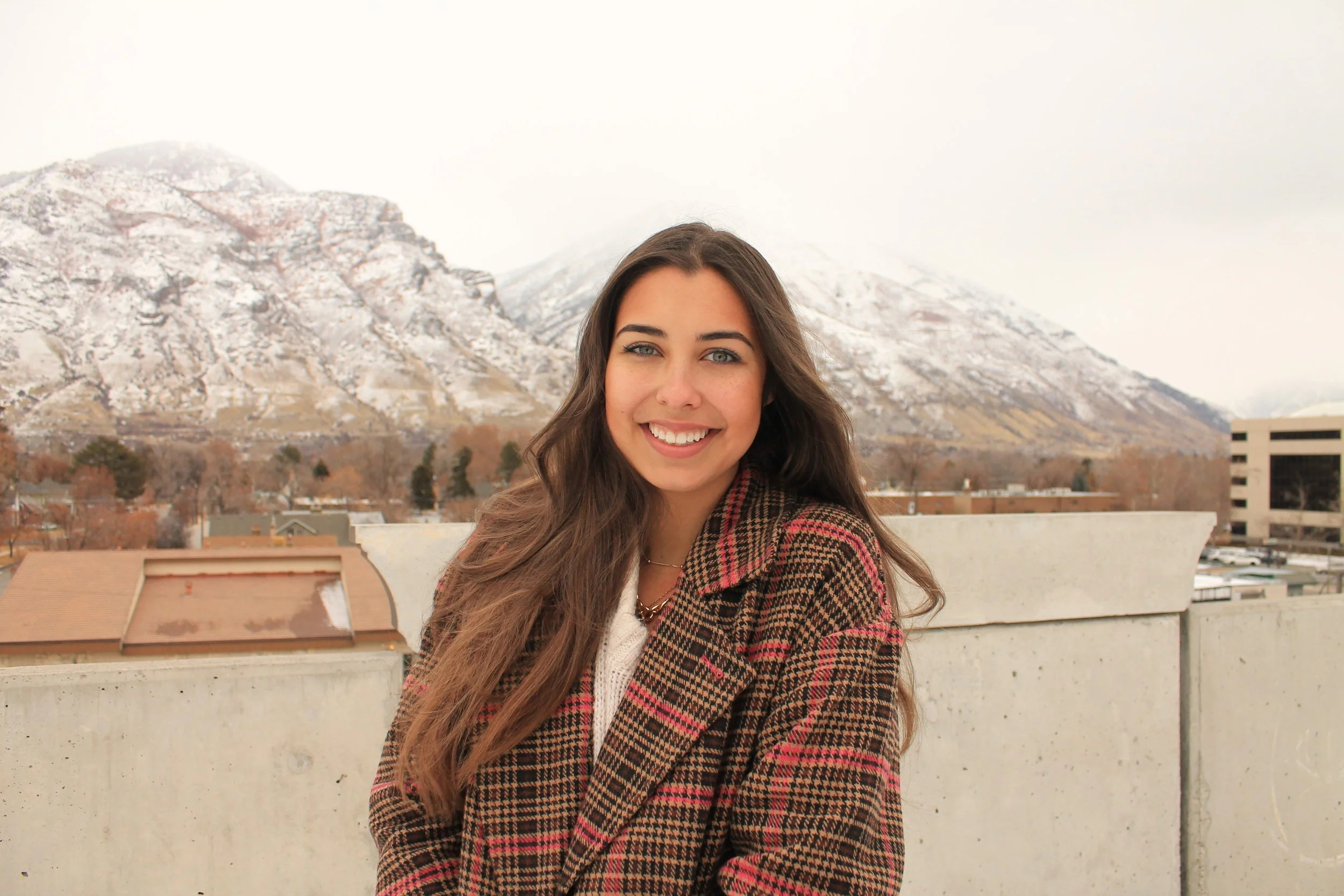 Young woman with long wavy brown hair smiling outdoors with a snow-capped mountain in the background.
