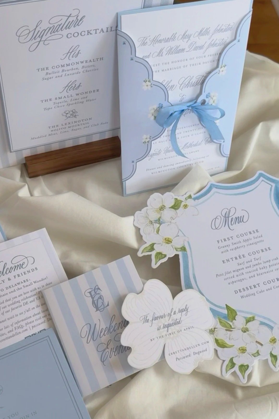 Wedding invitation suite with blue accents, floral decorations, and a menu card, displayed on ivory fabric.