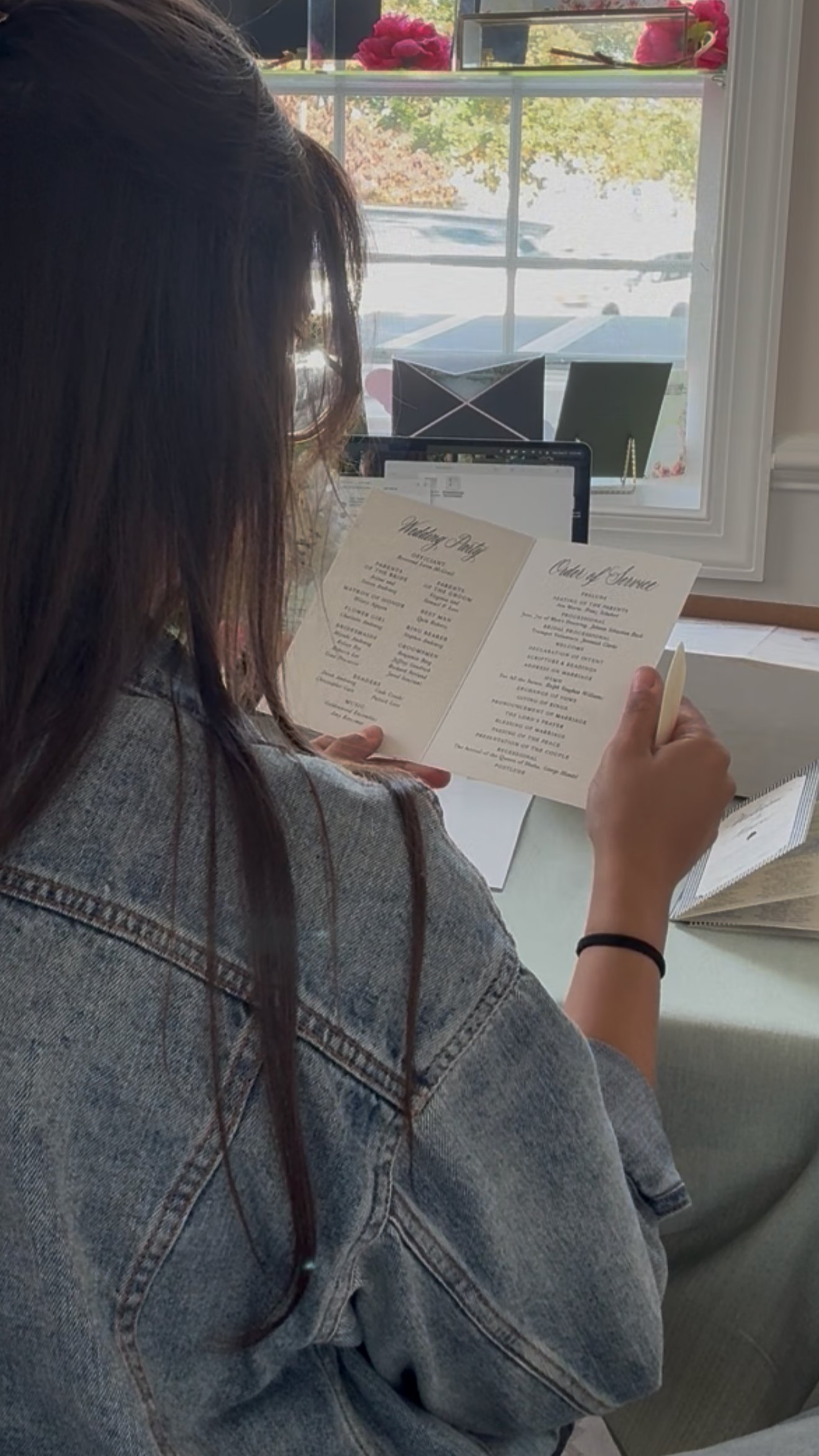 Person reading a wedding program at a table near a window with pink flowers on the windowsill.