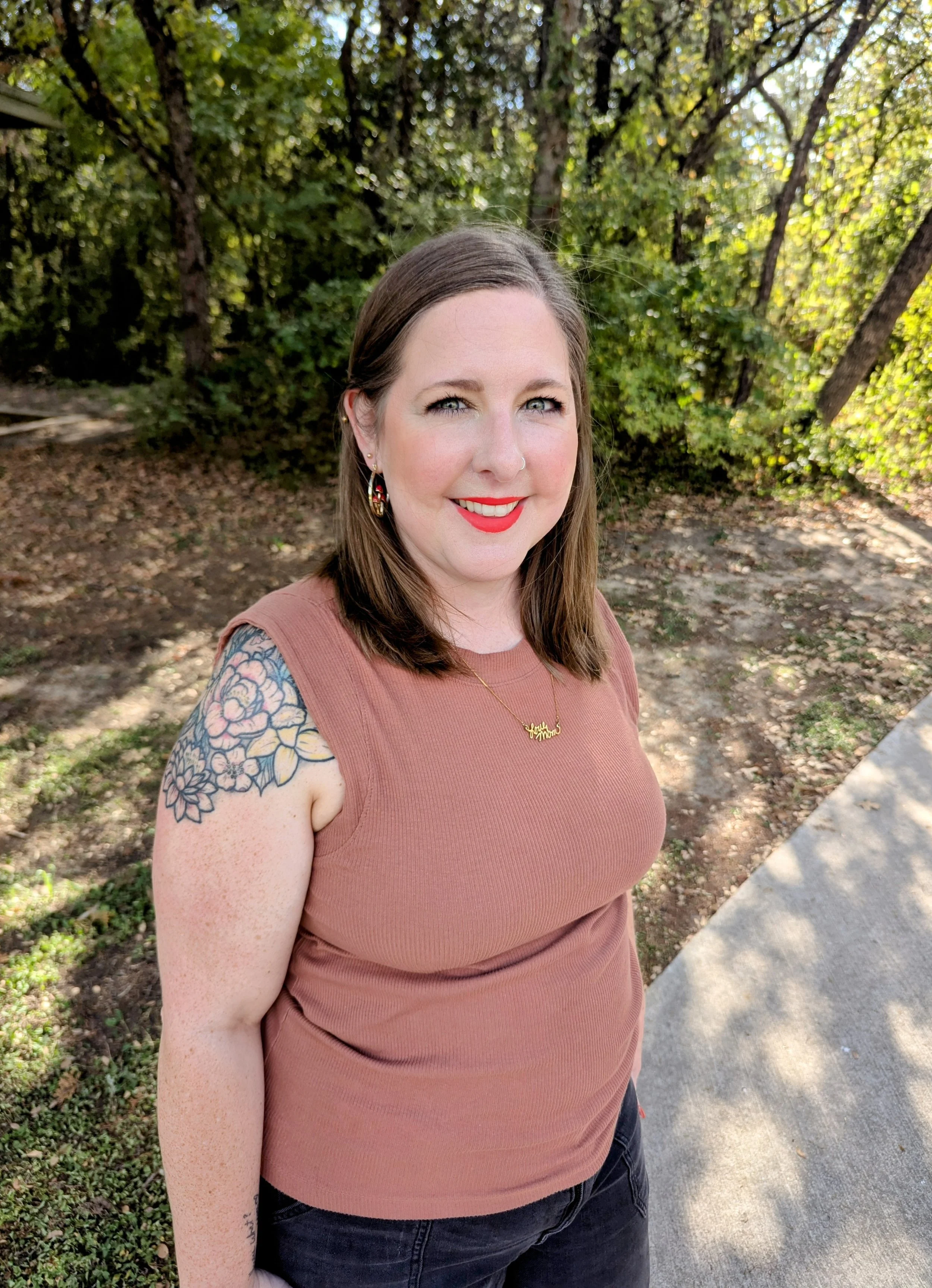 A woman smiling outdoors with a wooded background, wearing earrings, a nose ring, a necklace, and a short-sleeved pink top, showing a tattoo on her left arm.