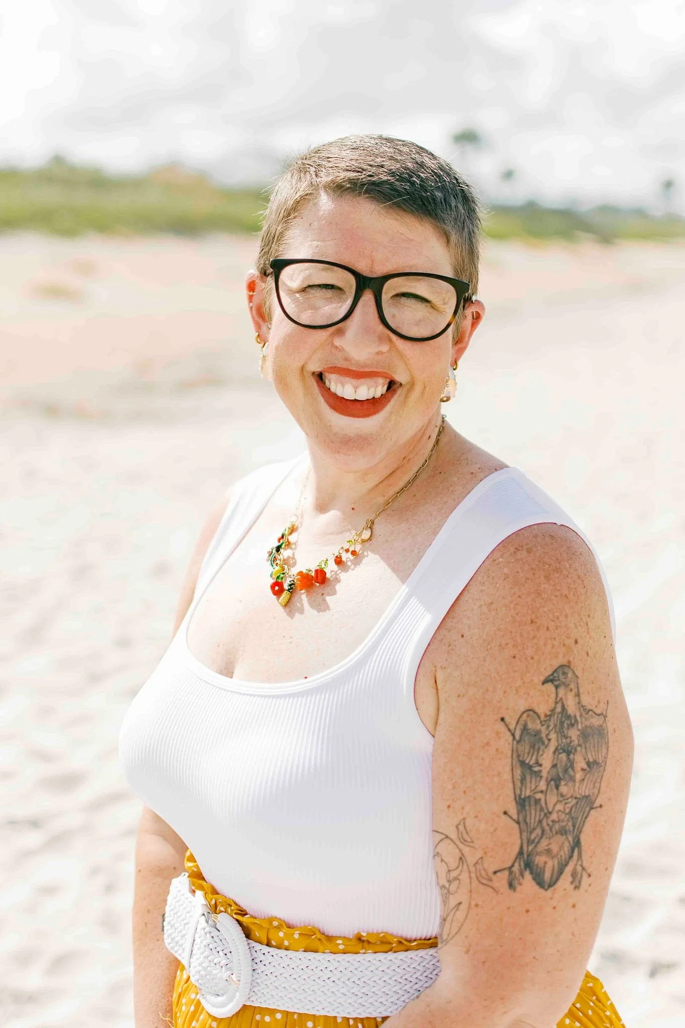 Close-up of a smiling woman with short gray hair, wearing black glasses, a white tank top, a colorful beaded necklace, and yellow polka dot shorts, standing on a beach with a blurred background of sand, grass, and sky.