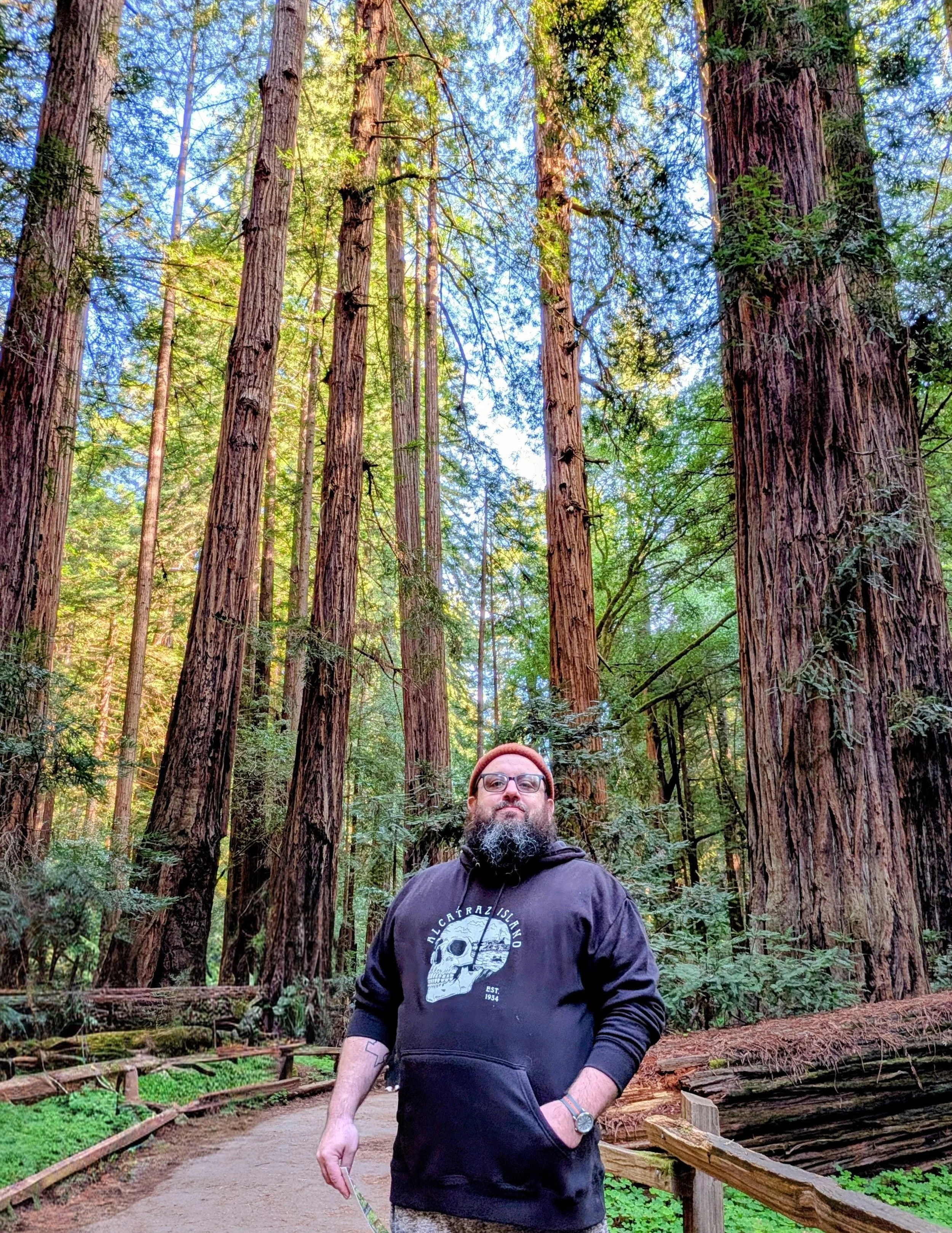 A man with glasses and a beard wearing a hoodie with a skull design stands on a forest trail surrounded by tall redwood trees in a lush green forest.